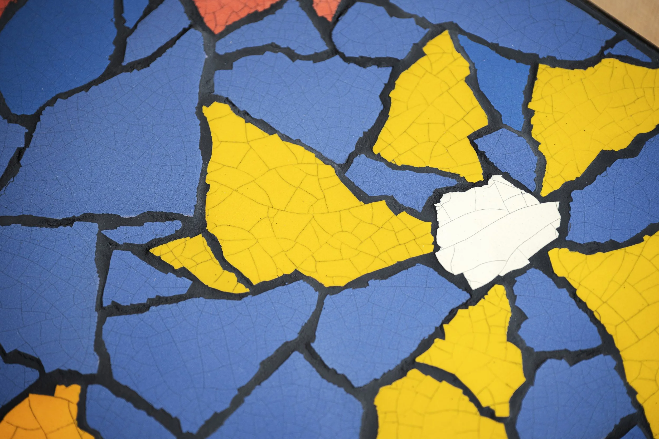 Close-up of a mosaic with irregularly shaped tiles in blue, yellow, white, and orange, separated by black grout lines.