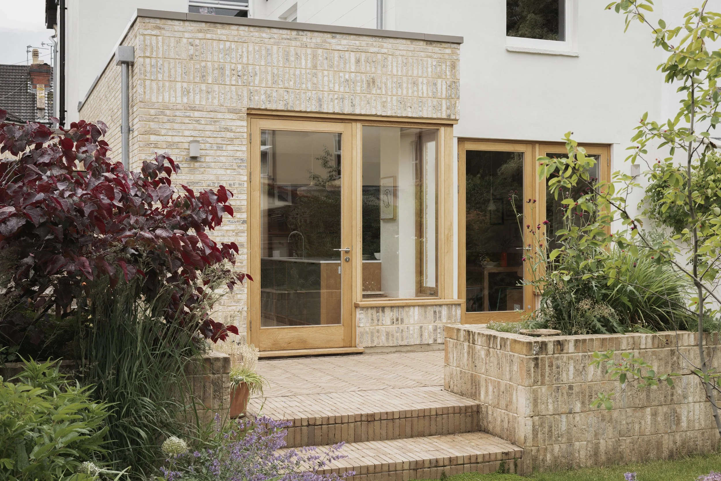 Exterior view of a modern house with a brick and white wall, large glass door, and window frames, surrounded by a garden with various plants and flowers.