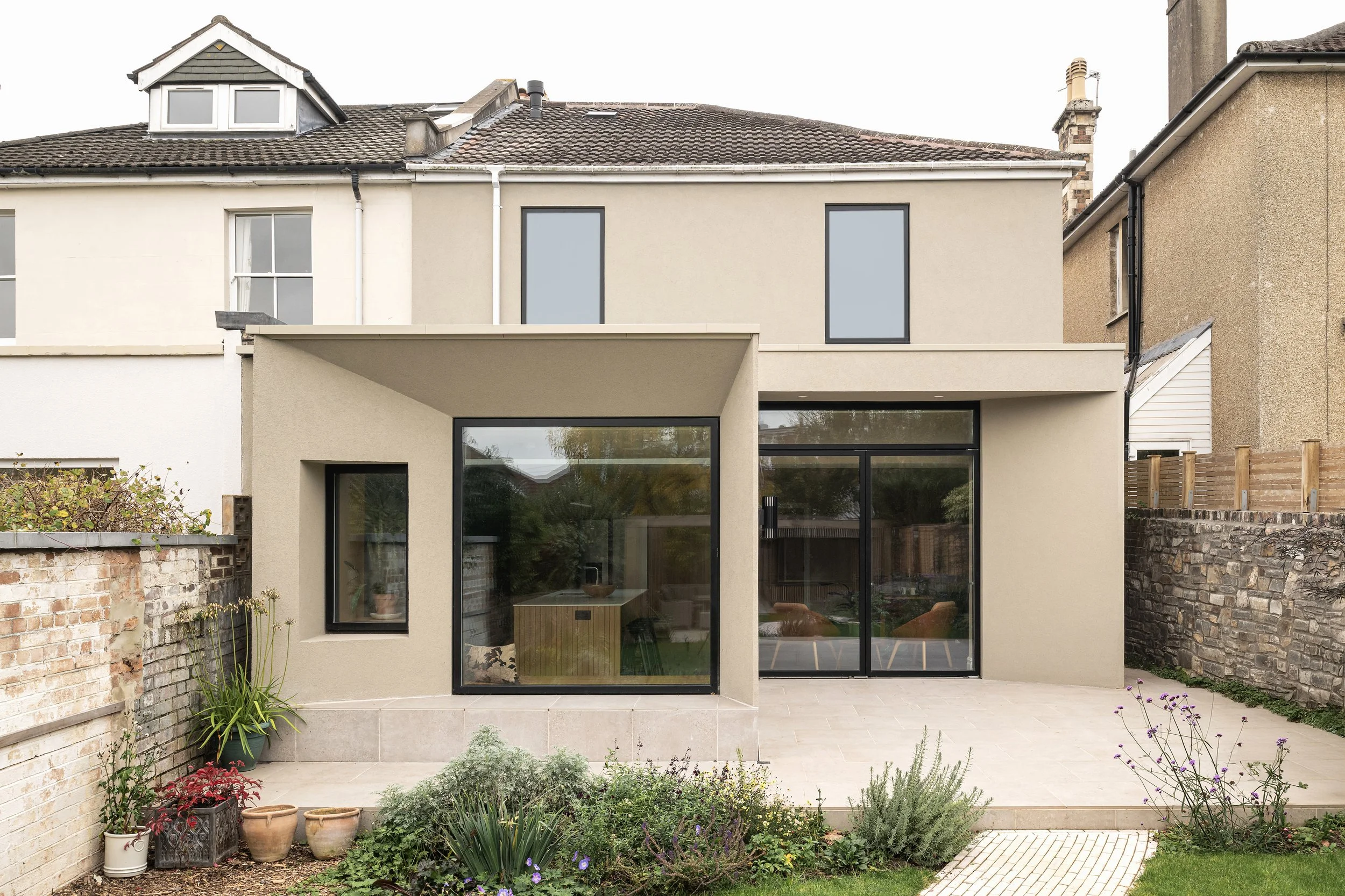 The rear view of a modern two-story house with large glass doors and windows, a beige exterior, a small garden with various plants and flowers, and a patio area.