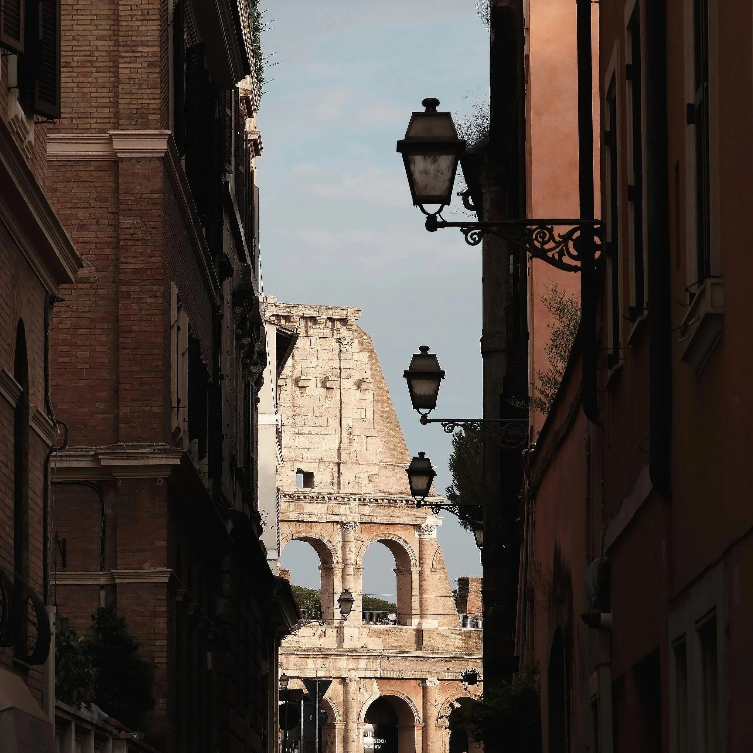 Narrow street with old buildings on both sides, featuring decorative architectural elements and vintage street lamps, leading to the Colosseum in the distance under a partly cloudy sky.