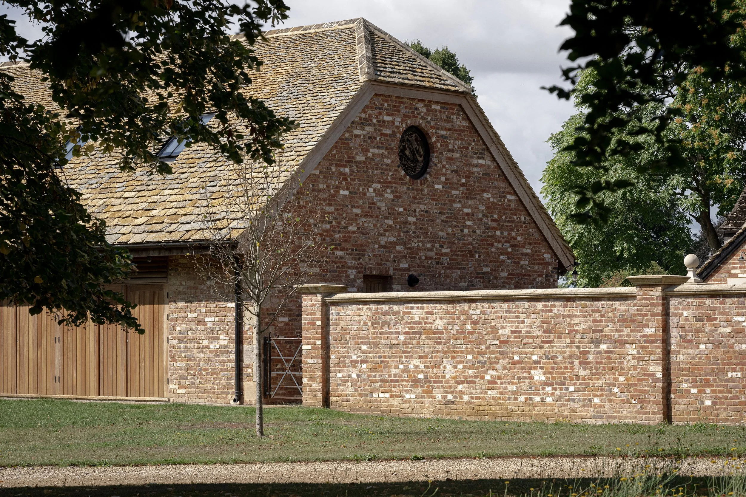 A brick building with a sloped tiled roof, a circular window near the roof's peak, and a brick wall in front with a wooden fence and a small tree in the yard.
