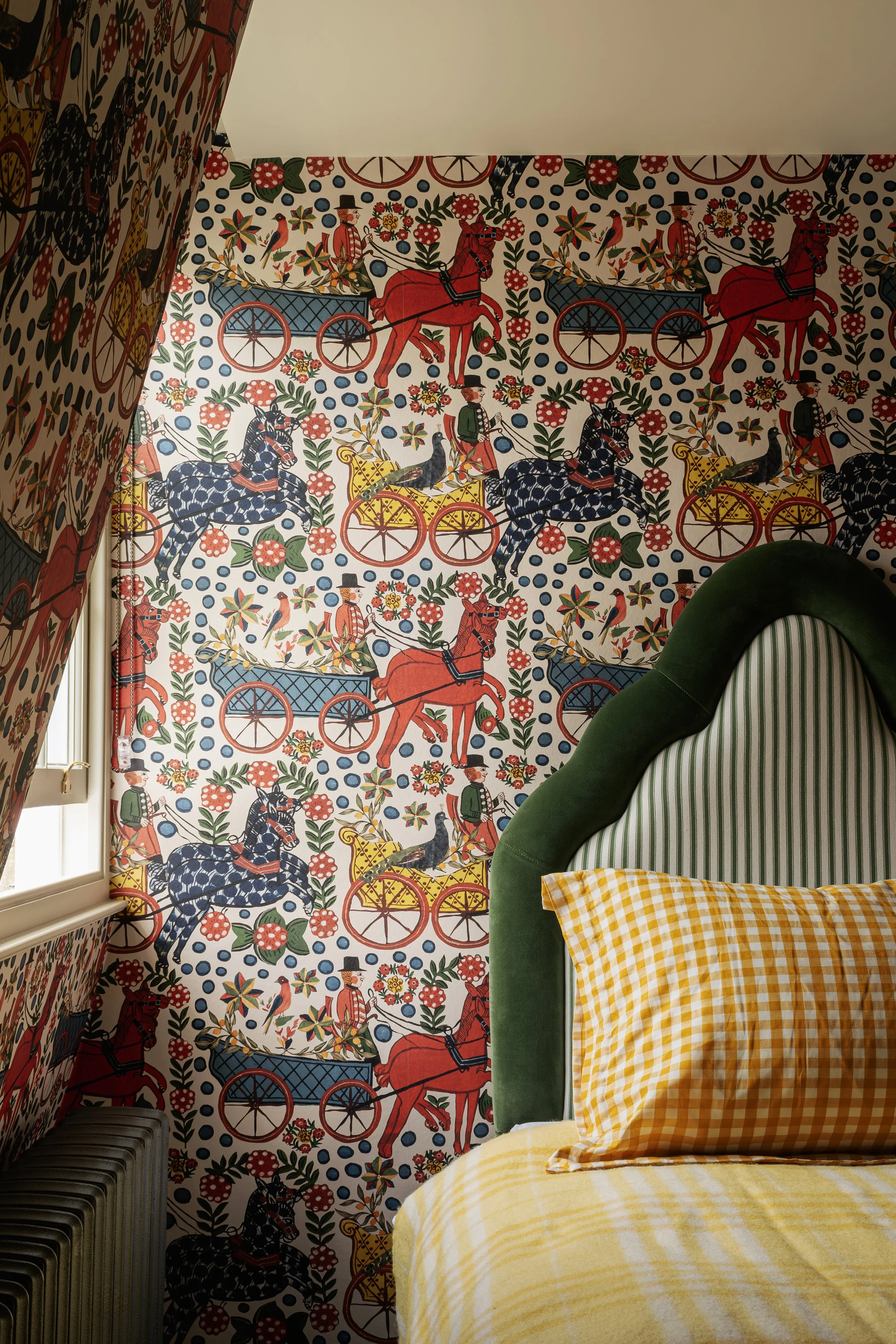 Colorful wallpaper with horse-drawn carriages, flowers, and people in traditional clothing, along with a green and striped headboard, a yellow checkered pillow, and a window with curtain on the left side.
