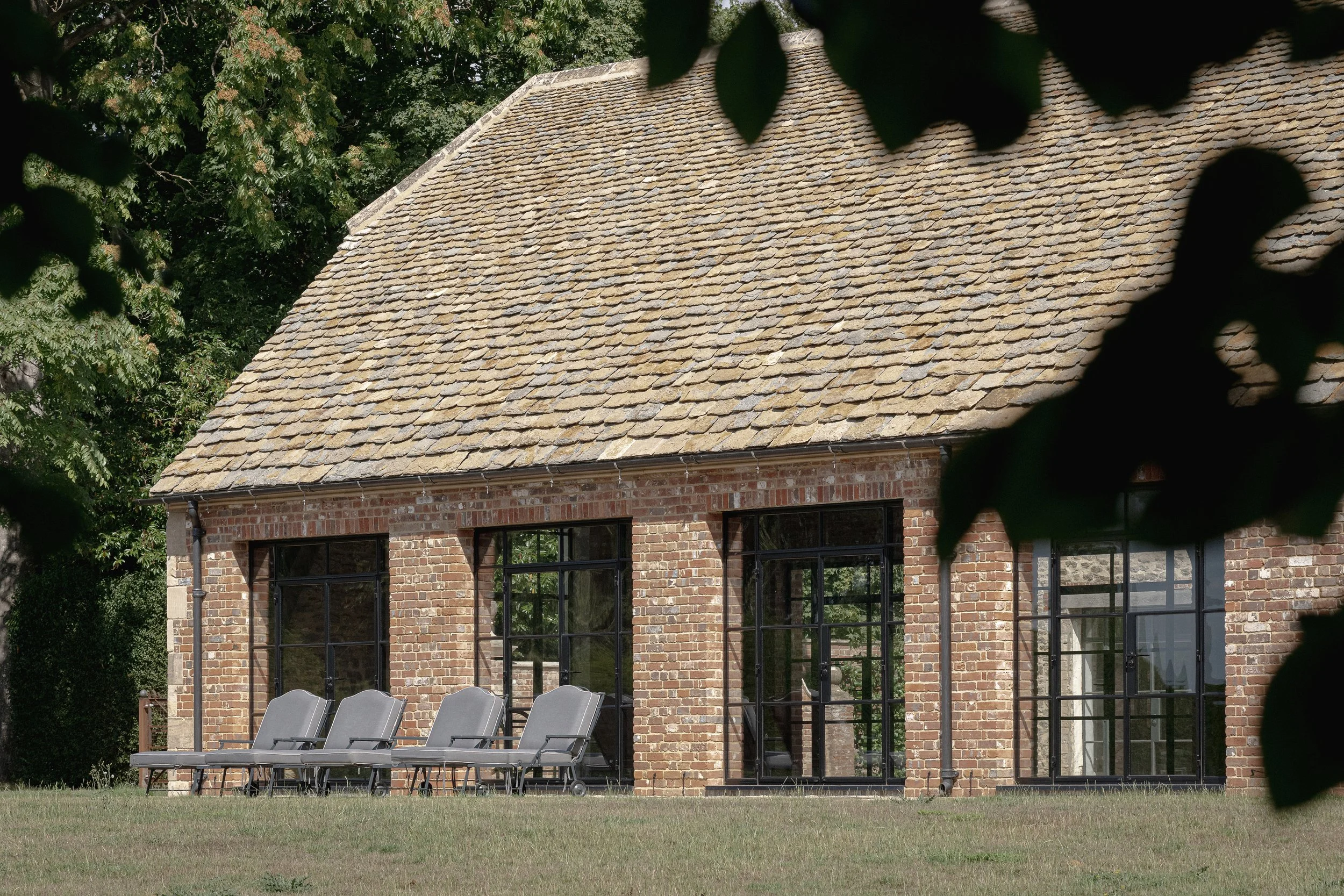 A brick house with large windows and a tiled roof, outdoor lounge chairs on the lawn in front, surrounded by trees.