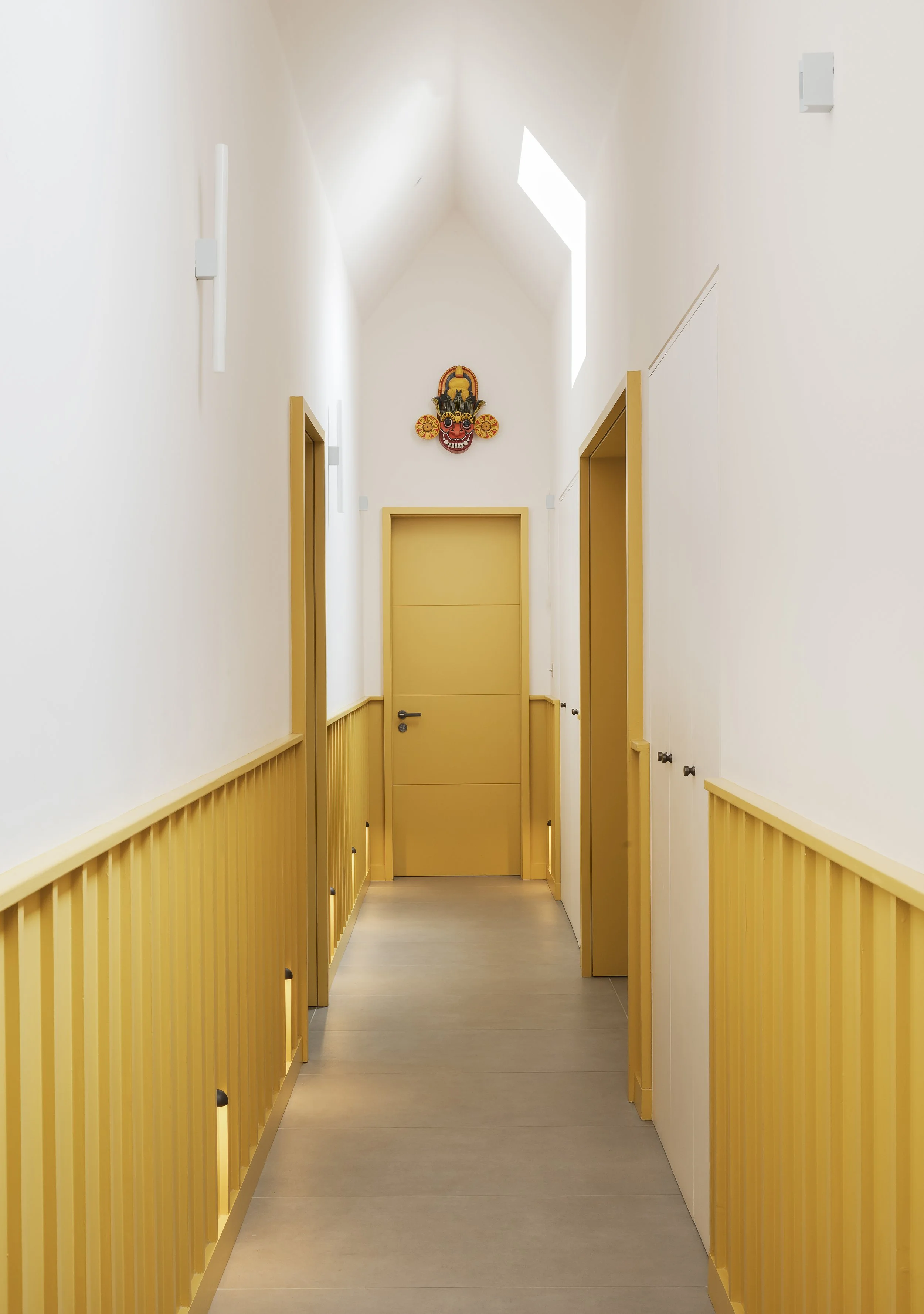 Bright hallway with white walls, yellow trim, and doors, featuring a colorful decorative mask at the end.