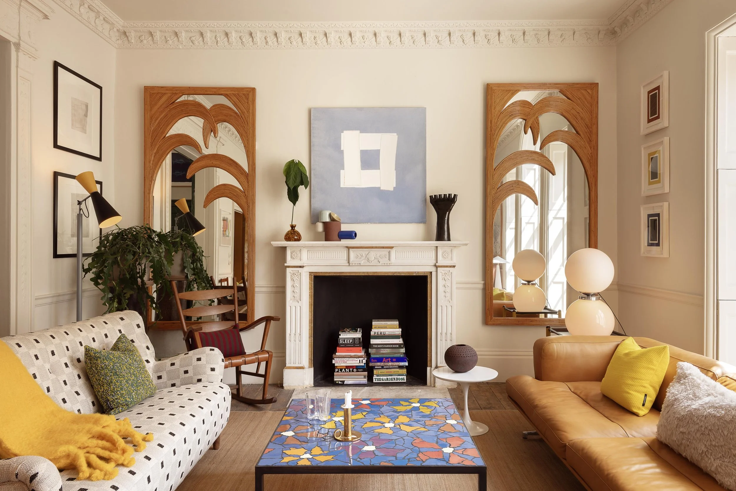 Living room with white walls and ornate crown molding, featuring a fireplace with books on the mantle and decorative objects, two large mirrors on either side of the fireplace, a patterned armchair with a yellow throw, a tan leather sofa with yellow 