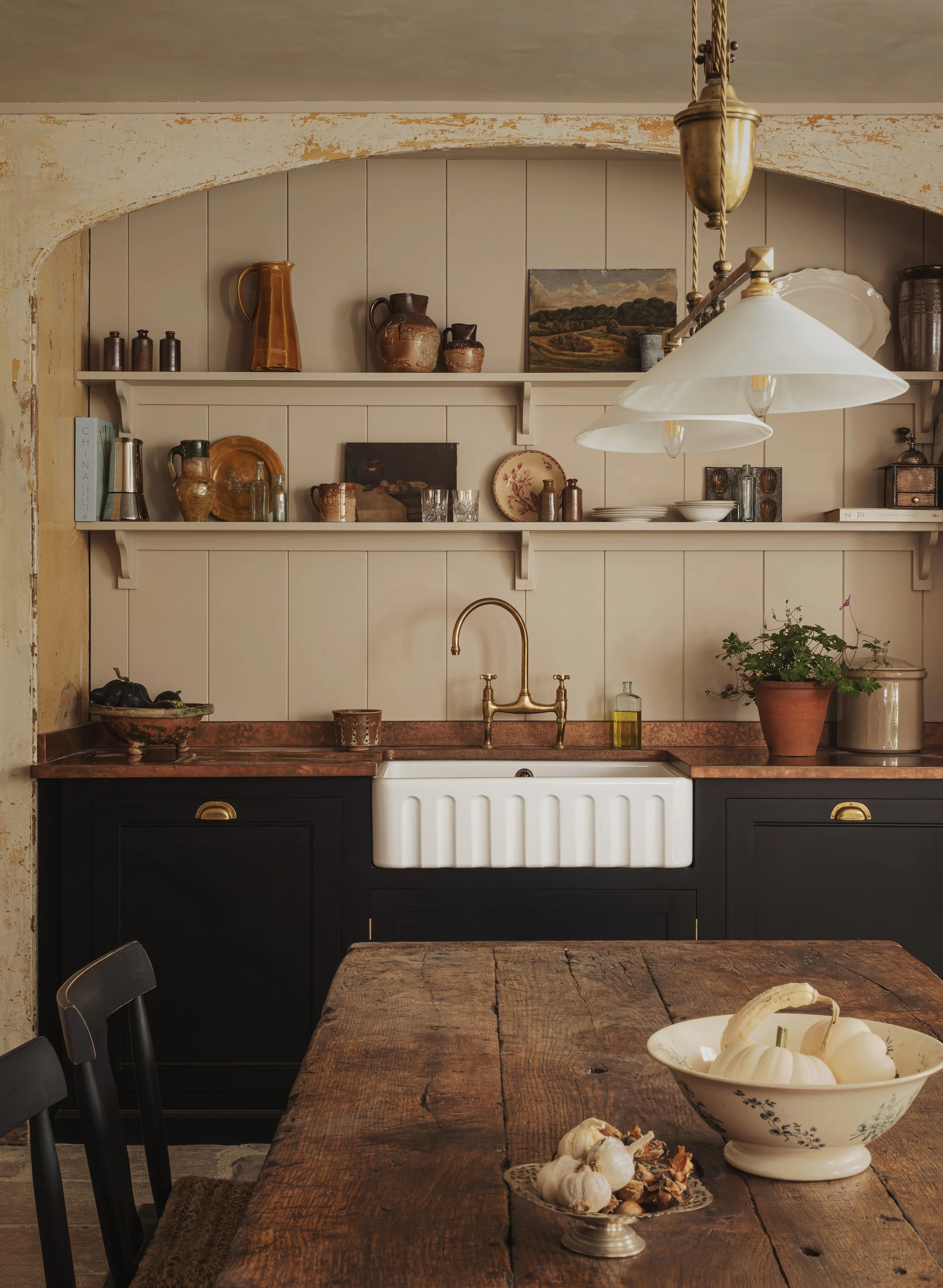 Kitchen with rustic wooden table, white farmhouse sink, black lower cabinets, beige wall with open shelves holding pottery, books, and decorative items, pendant lights hanging above, potted plant on countertop.