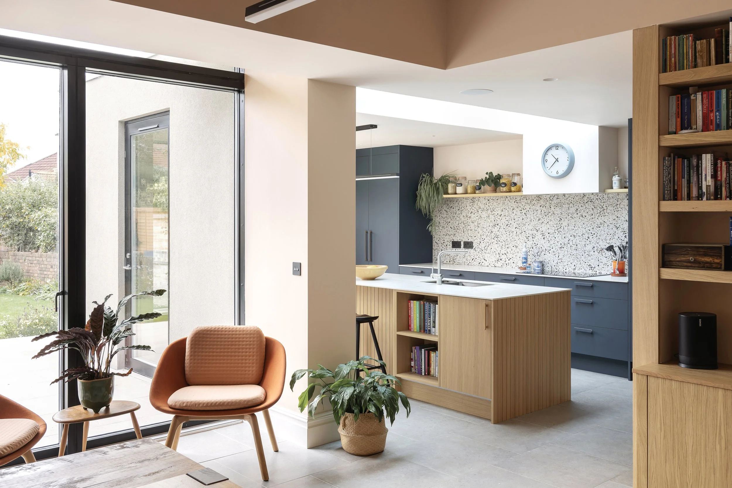 Modern open-plan kitchen and living area with orange chairs, potted plants, blue and wood cabinetry, and a terrazzo backsplash.