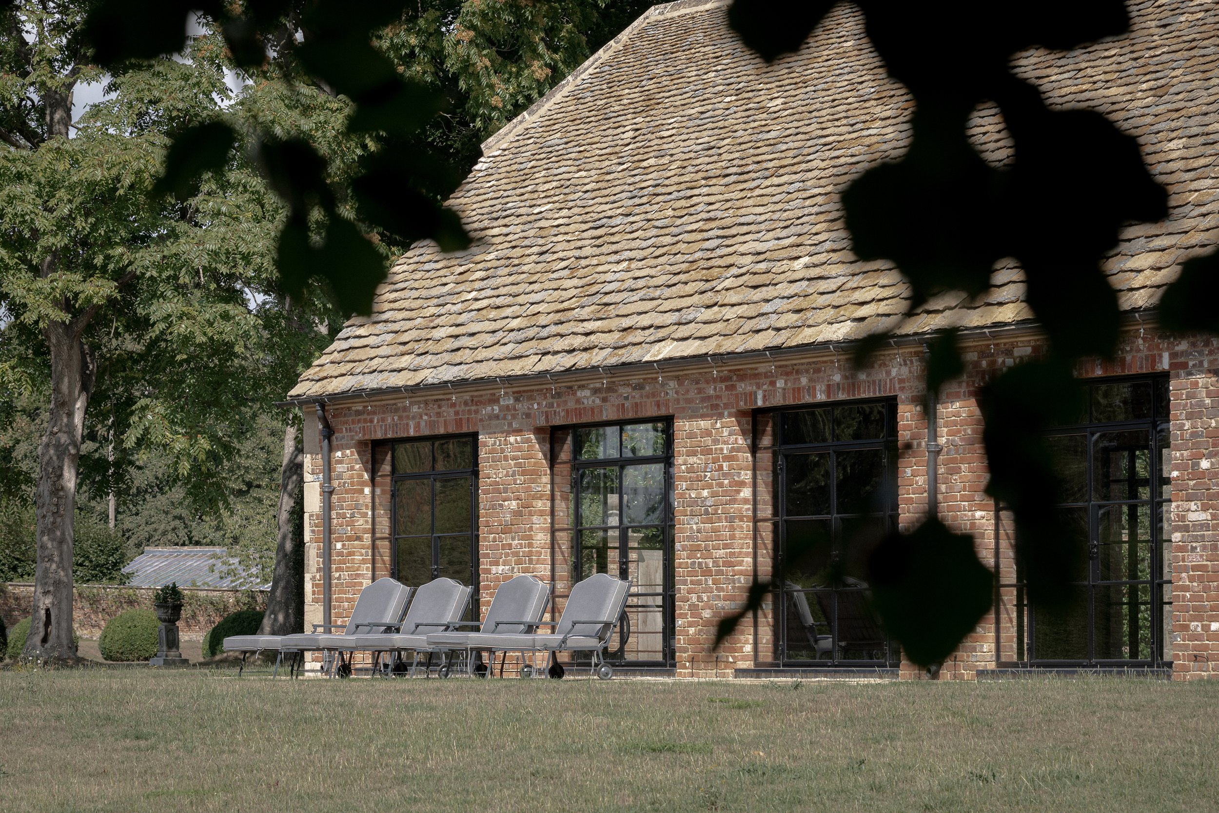 A brick building with large black-framed windows and a shingled roof, partially obscured by tree branches and leaves in the foreground.