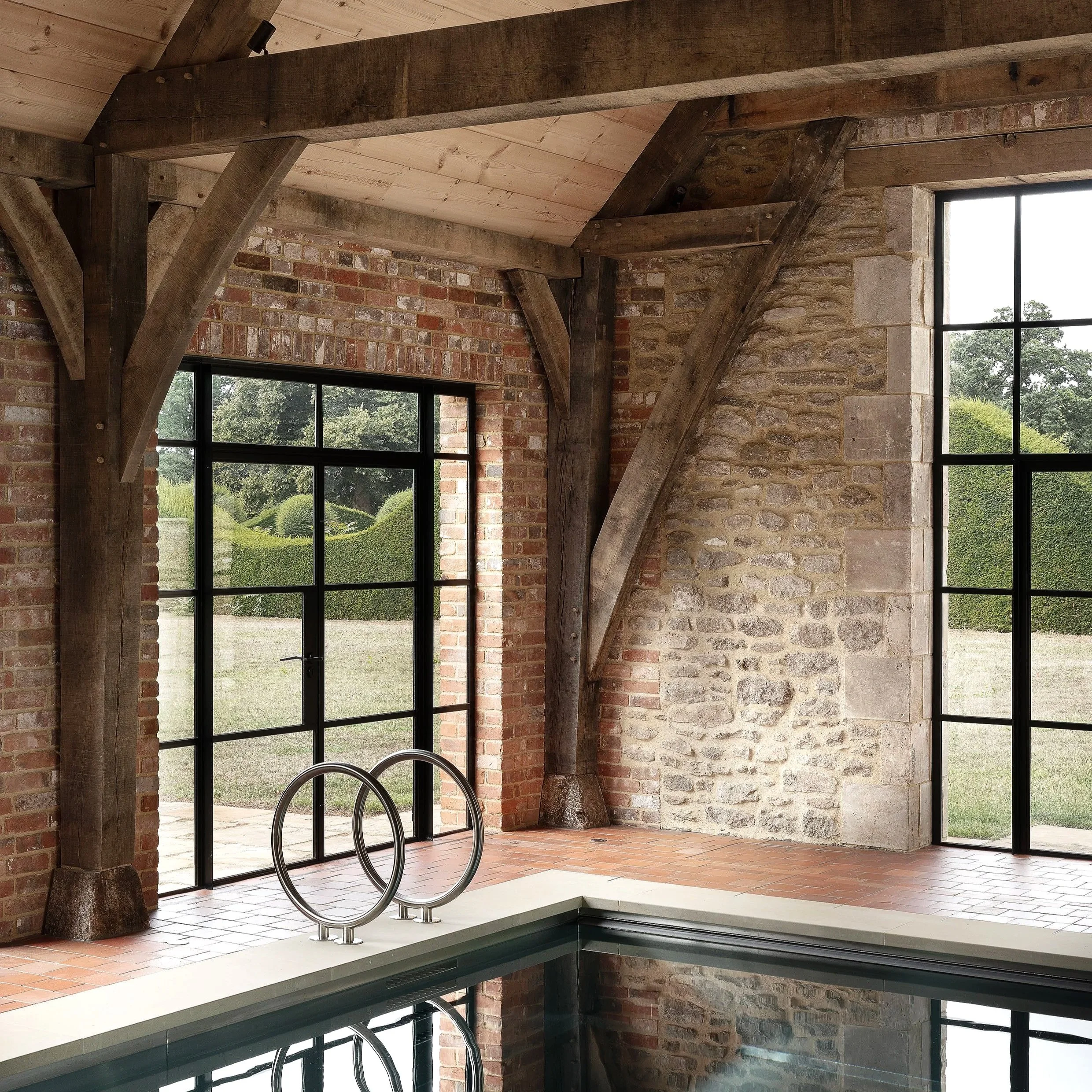 Indoor swimming pool area with large windows, exposed brick and stone walls, wooden beams, and a metal pool ladder.