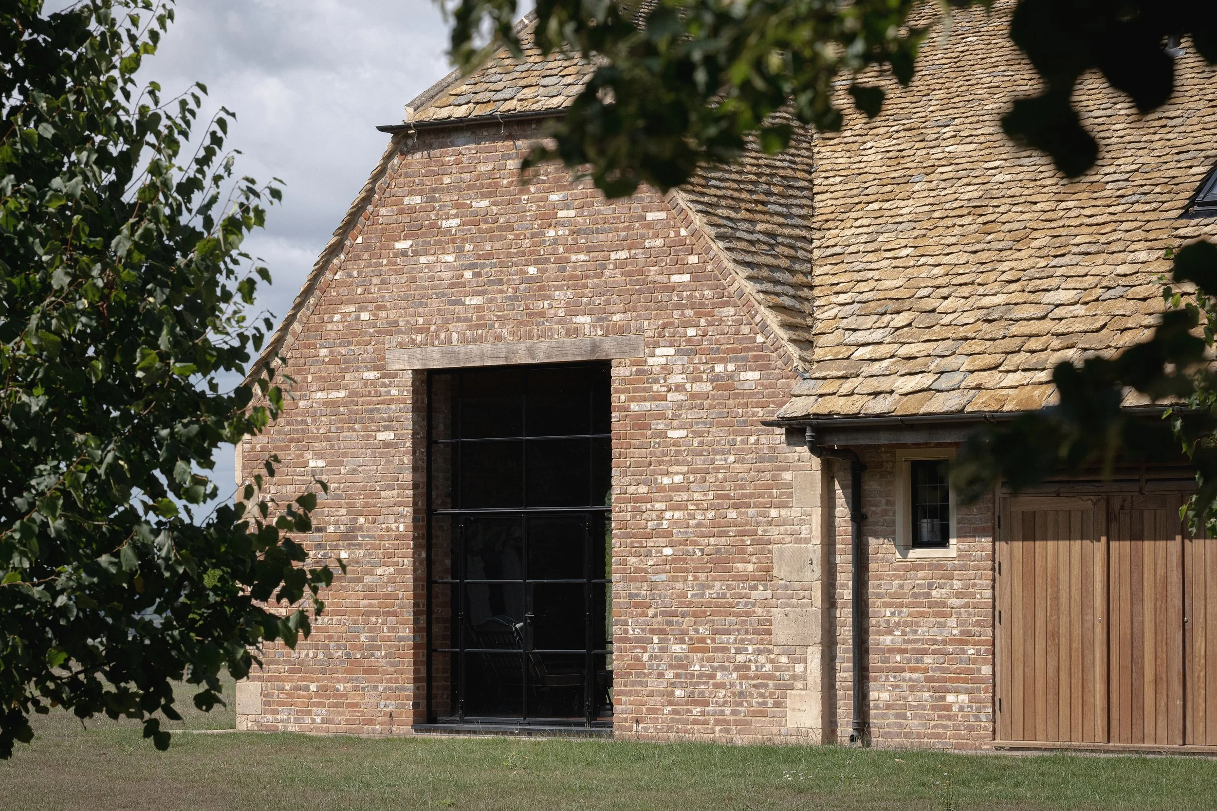 A brick building with a large black metal door, small window, and a roof with brown shingles, partially obscured by green leaves in the foreground.