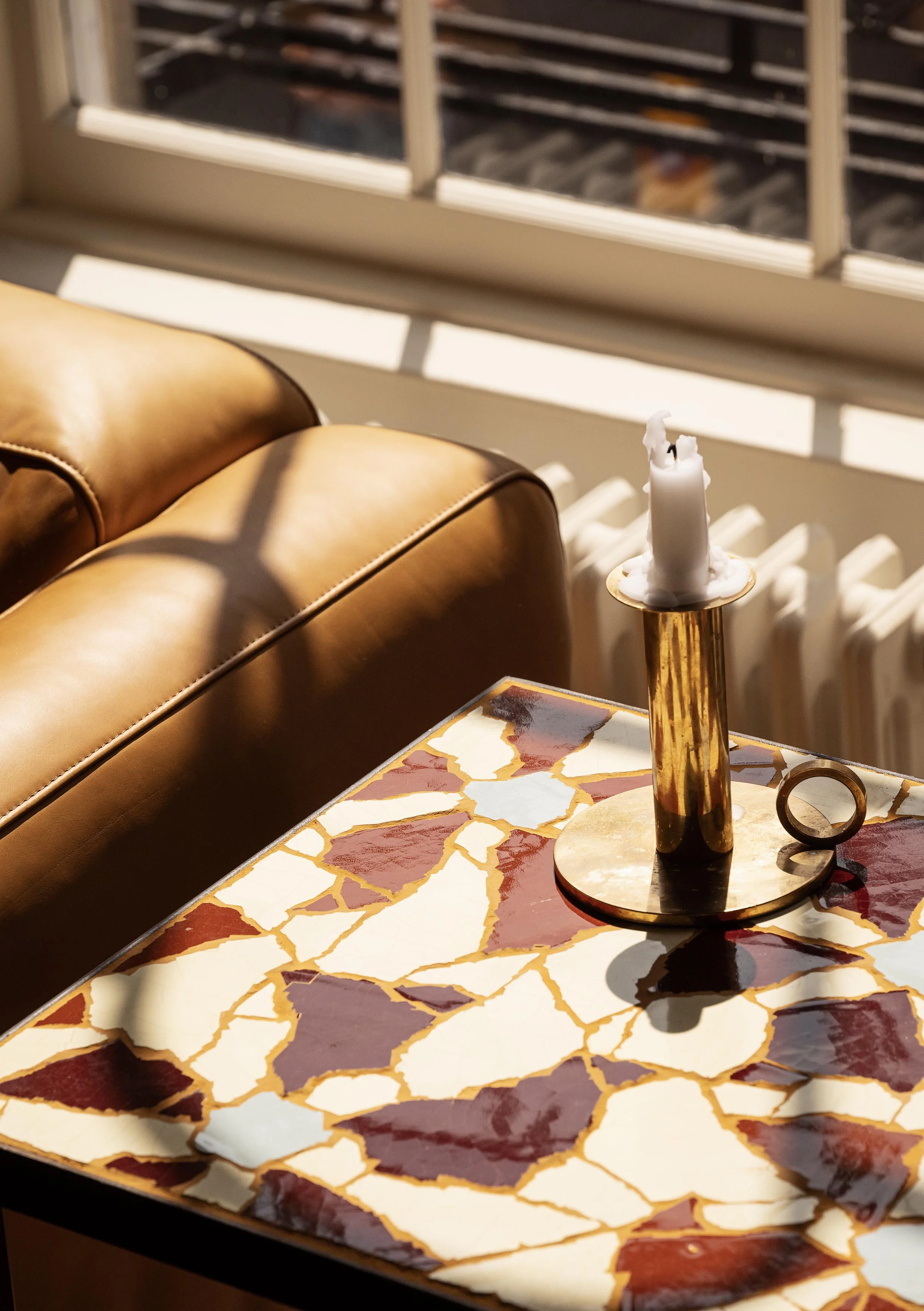 A close-up of a patterned table with a gold candle holder and a white, partially melted candle, with a leather couch and window in the background.