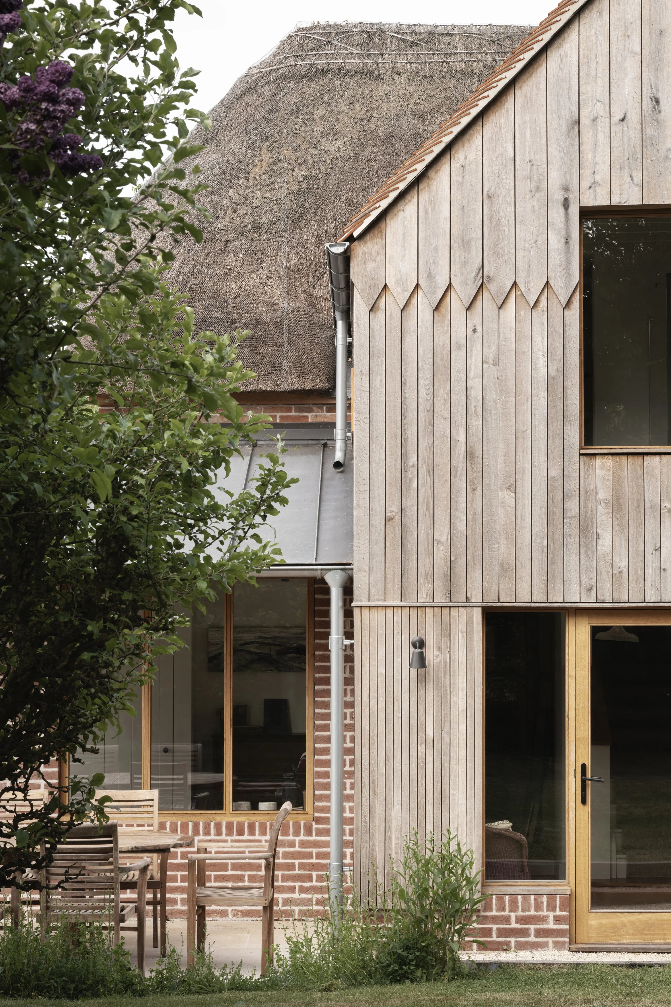 Front view of a house with a thatched roof on the left and vertical wooden siding on the right, with a large window and glass door, a small outdoor seating area with a table and four chairs, and some greenery in the foreground.