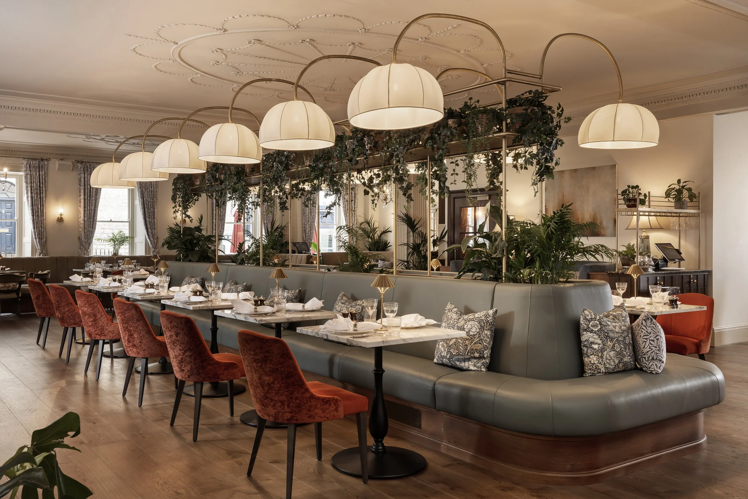 Stylish restaurant interior with a long table set for dining, modern pendant lights hanging overhead, a curved banquette seating with decorative pillows, and lush greenery in the background.