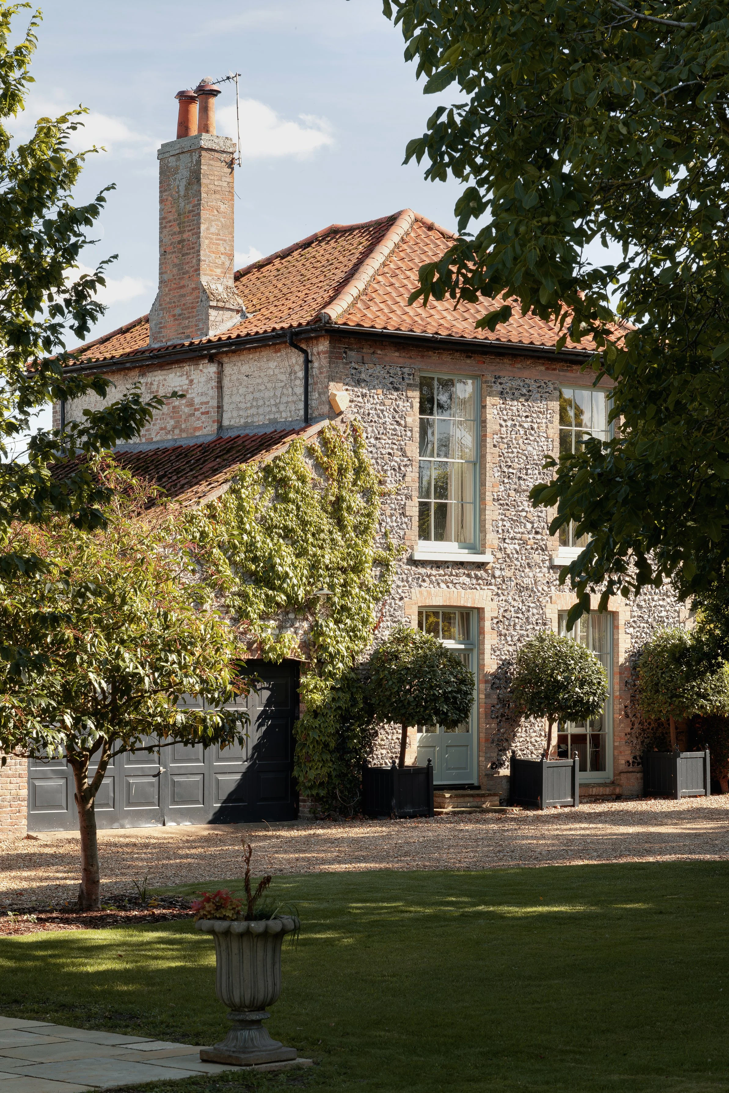 A two-story brick house with large windows, a red tiled roof, and a chimney, surrounded by greenery and a manicured lawn.