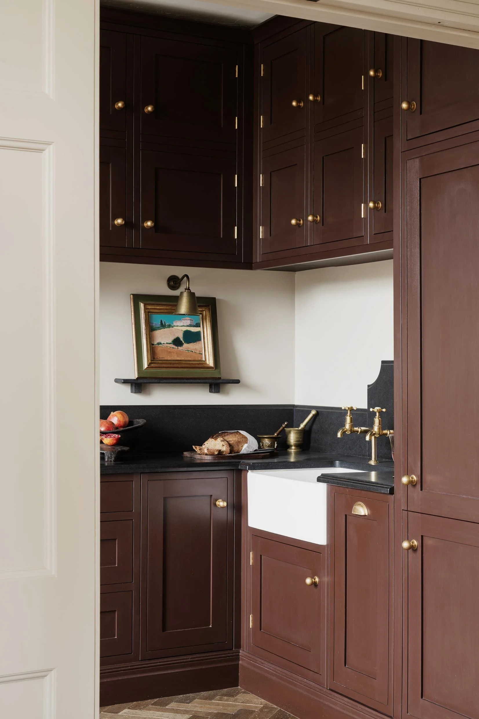 Kitchen with dark wood cabinets, gold knobs, a black countertop, and a white farmhouse sink, with apples and bread on the counter, a small shelf with a landscape painting, and a brass wall-mounted lamp above the painting.