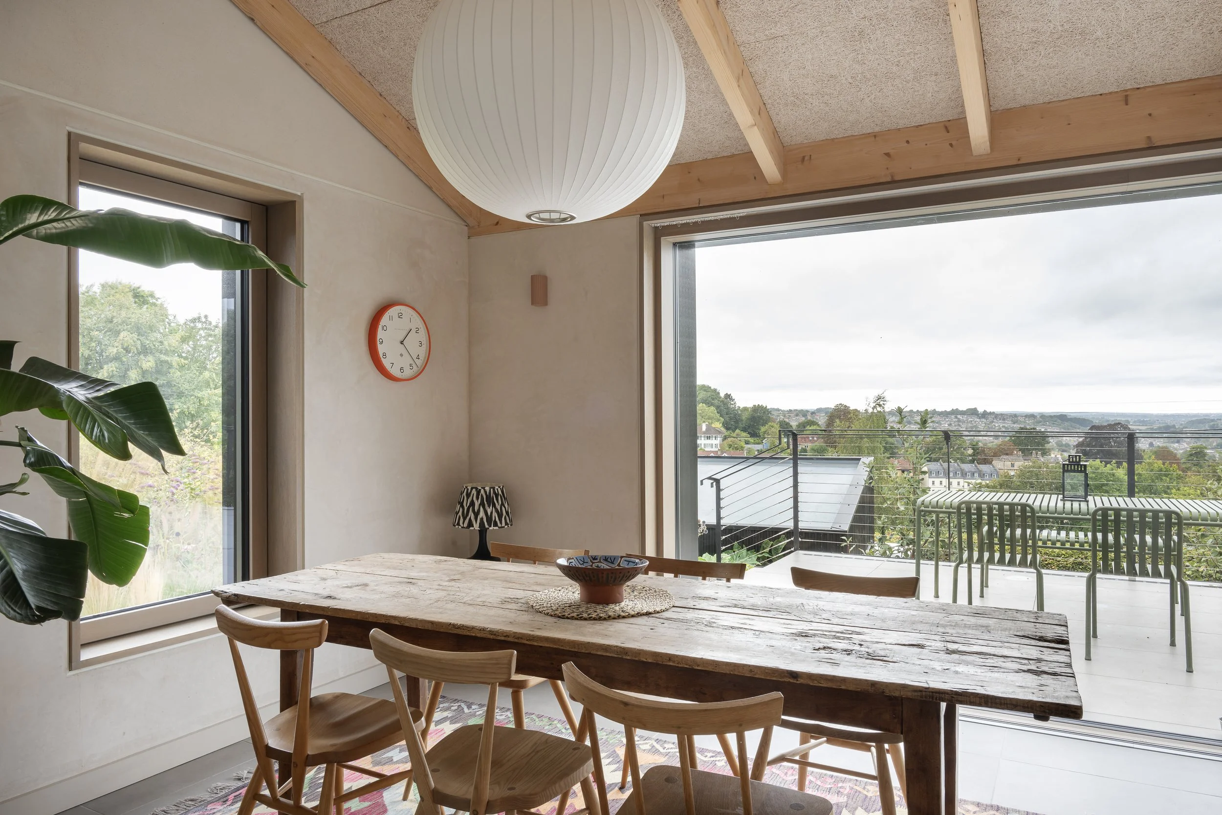 Inside a dining room with a rustic wooden table, six wooden chairs, a black and white patterned table lamp, a large round paper lantern, a clock showing 2:15, a large window with a view of a balcony and a semi-urban landscape with trees and rooftops.