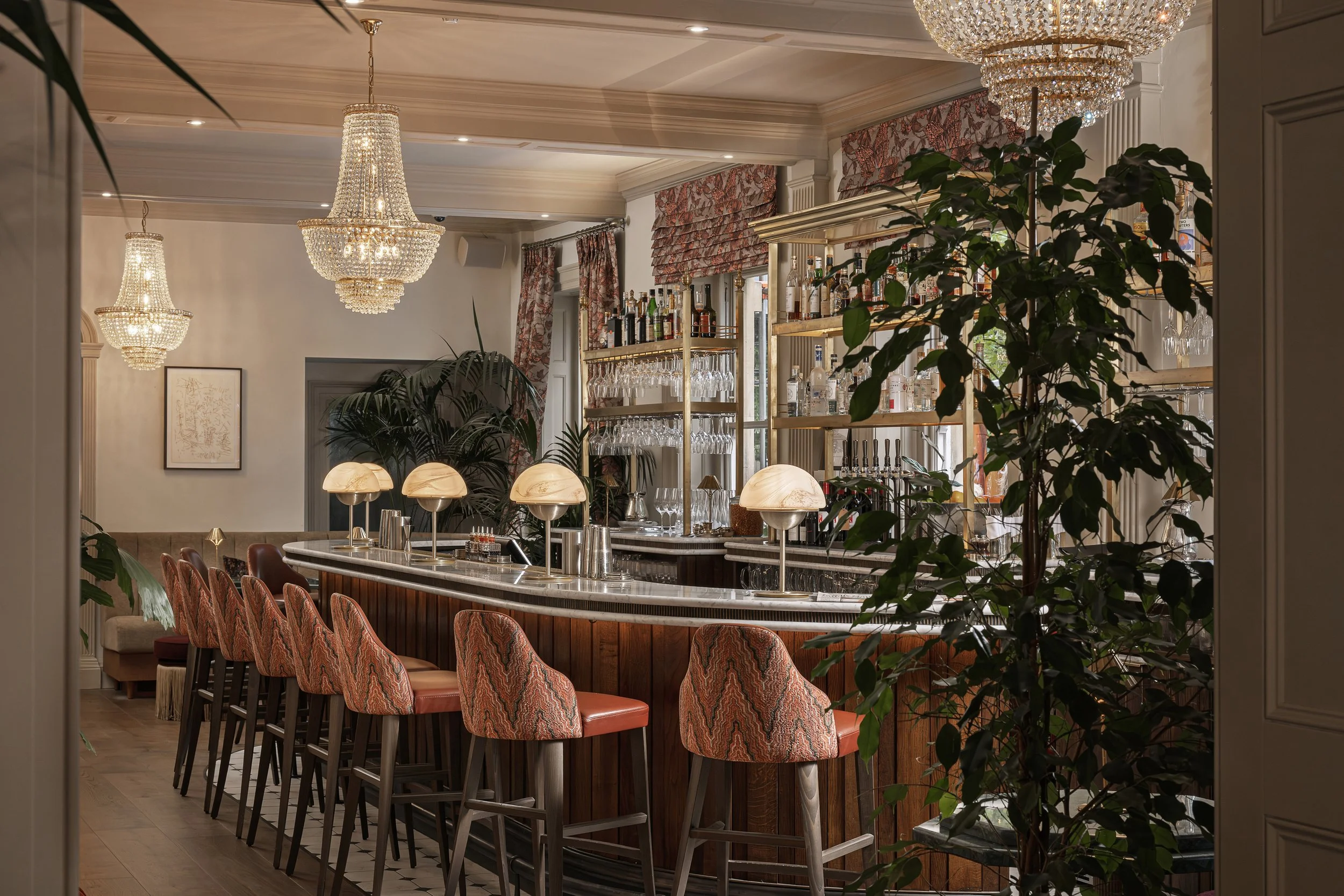 Elegant bar with a marble countertop, surrounded by patterned upholstered bar stools. Behind the bar, shelves display various bottles and glassware, with large windows and floral curtains. Crystal chandeliers hang from the ceiling, adding a touch of 