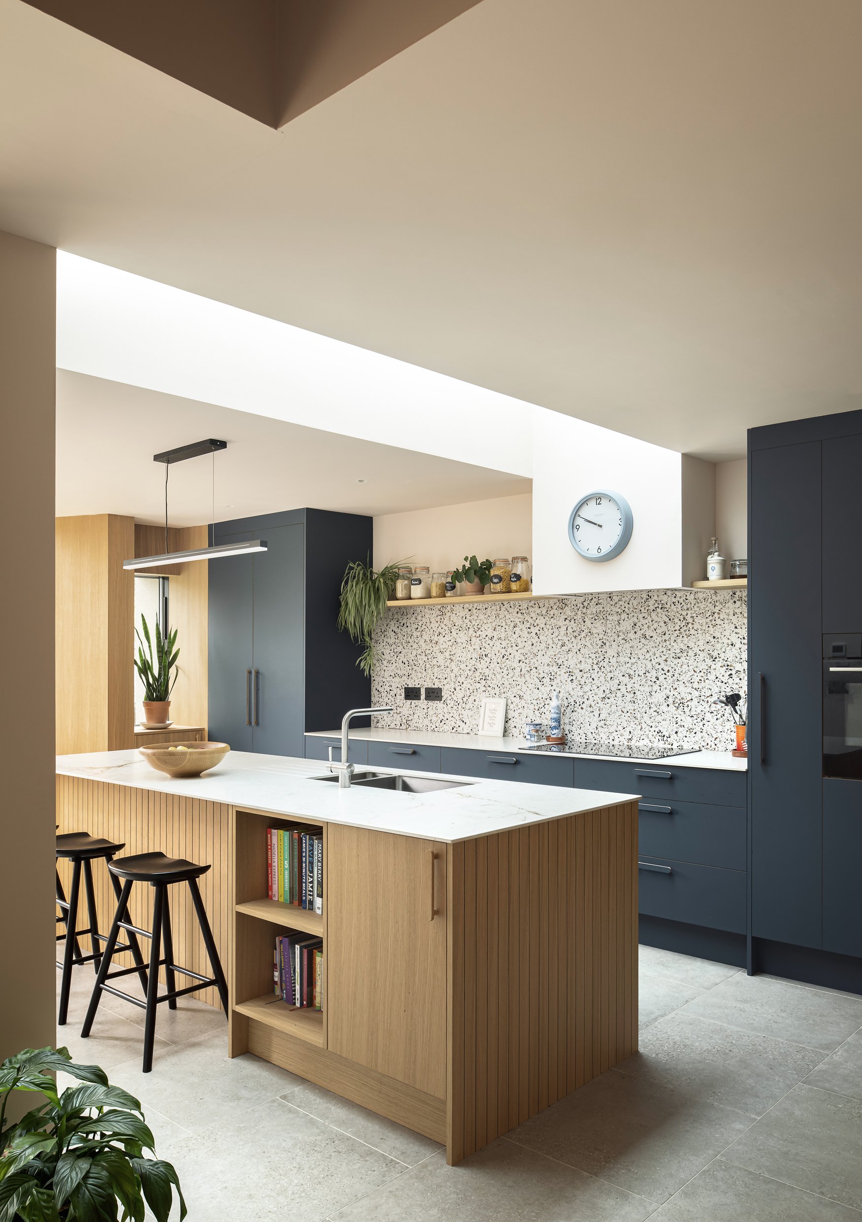 Modern kitchen with a white marble island, blue cabinets, and wooden accents. Potted plants are visible, along with a wall clock and some jars on a shelf.