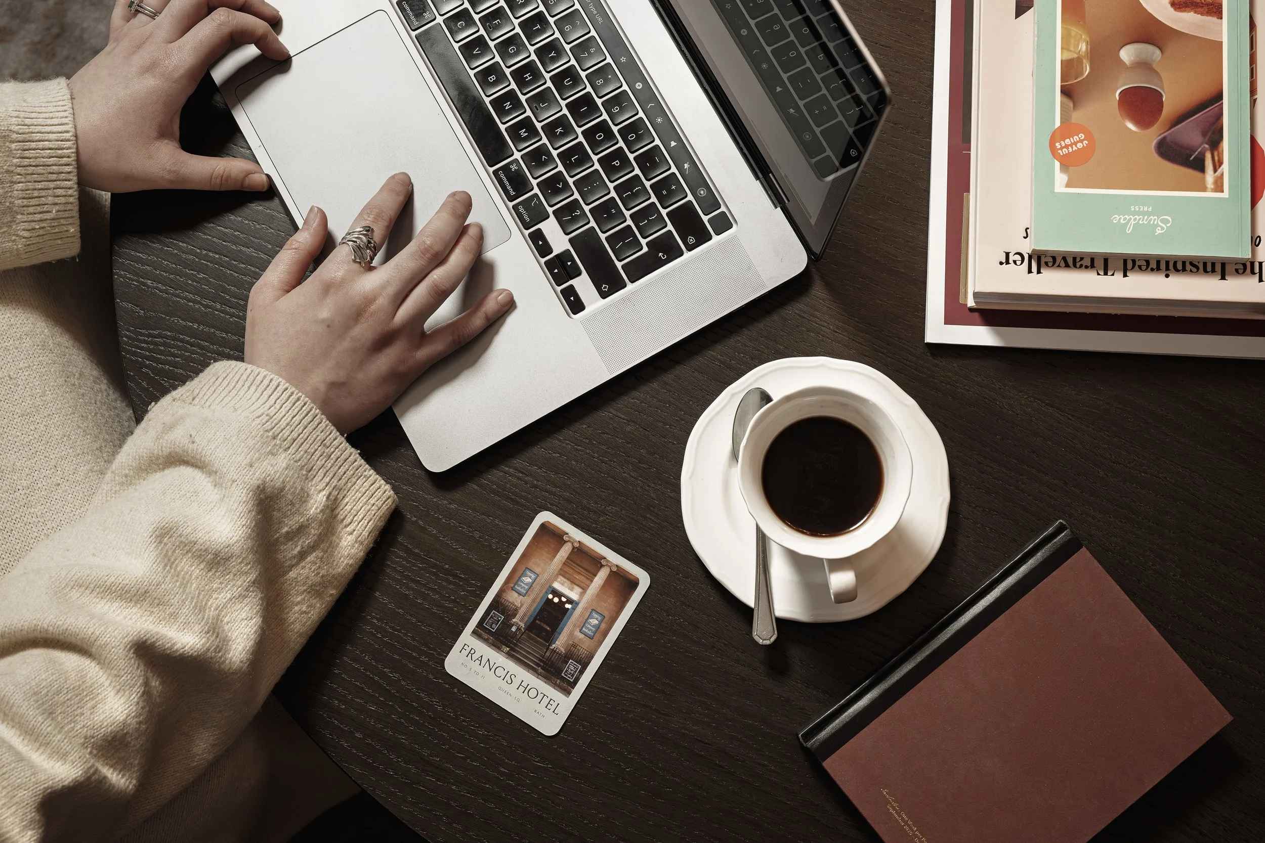 Person working on a laptop at a dark wooden table with a cup of coffee, a hotel keycard, books, and a colorful box on top of the books.