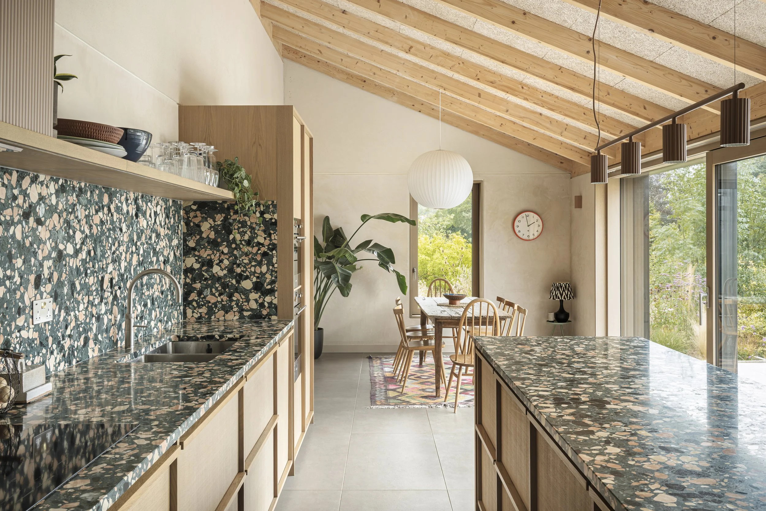 Open-concept kitchen with terrazzo countertops and wooden cabinetry, adjacent to a dining area with a wooden table, chairs, and large windows with views of greenery.
