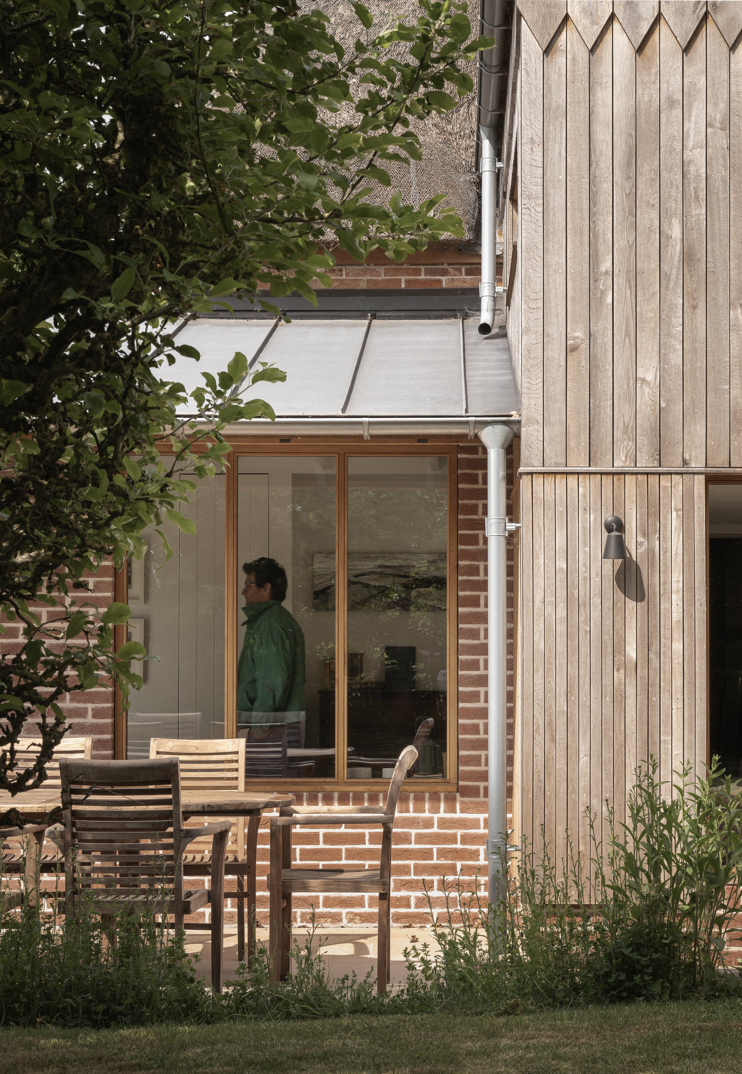 A person standing inside a house seen through large glass window, with a brick wall and wooden exterior walls, outdoor patio chairs, and green plants.