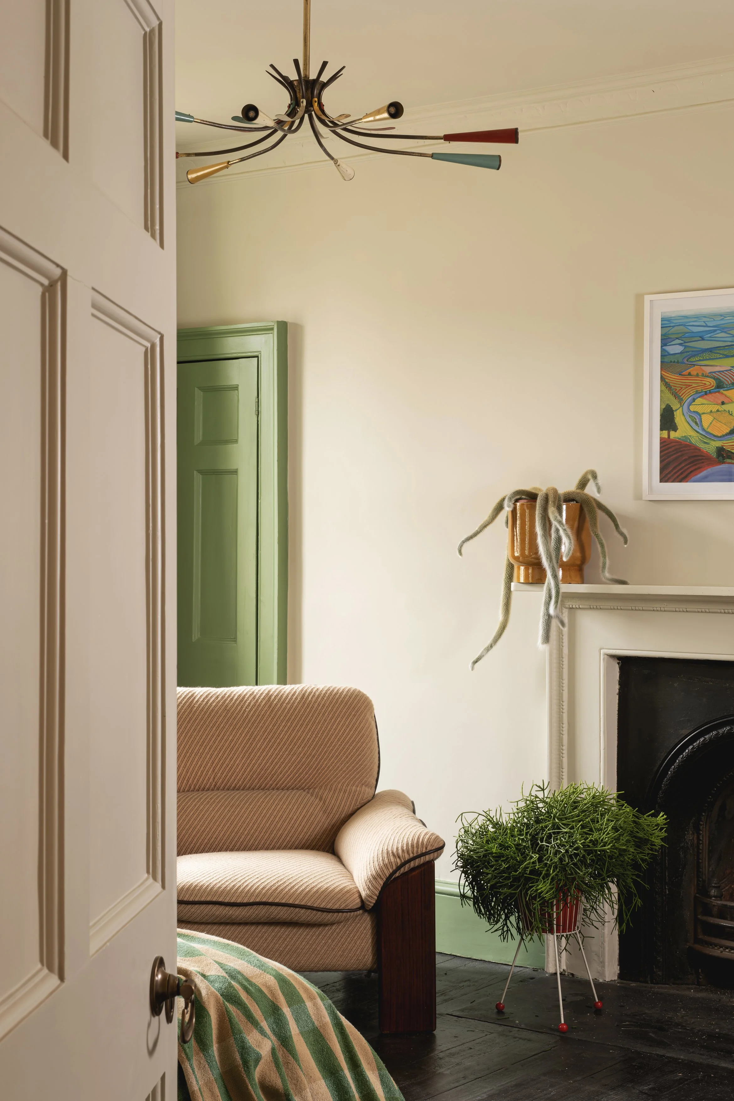 A cozy living room corner with a beige sofa, a potted plant on a stand, a colorful abstract painting, a green door, and a unique multicolored ceiling light fixture.