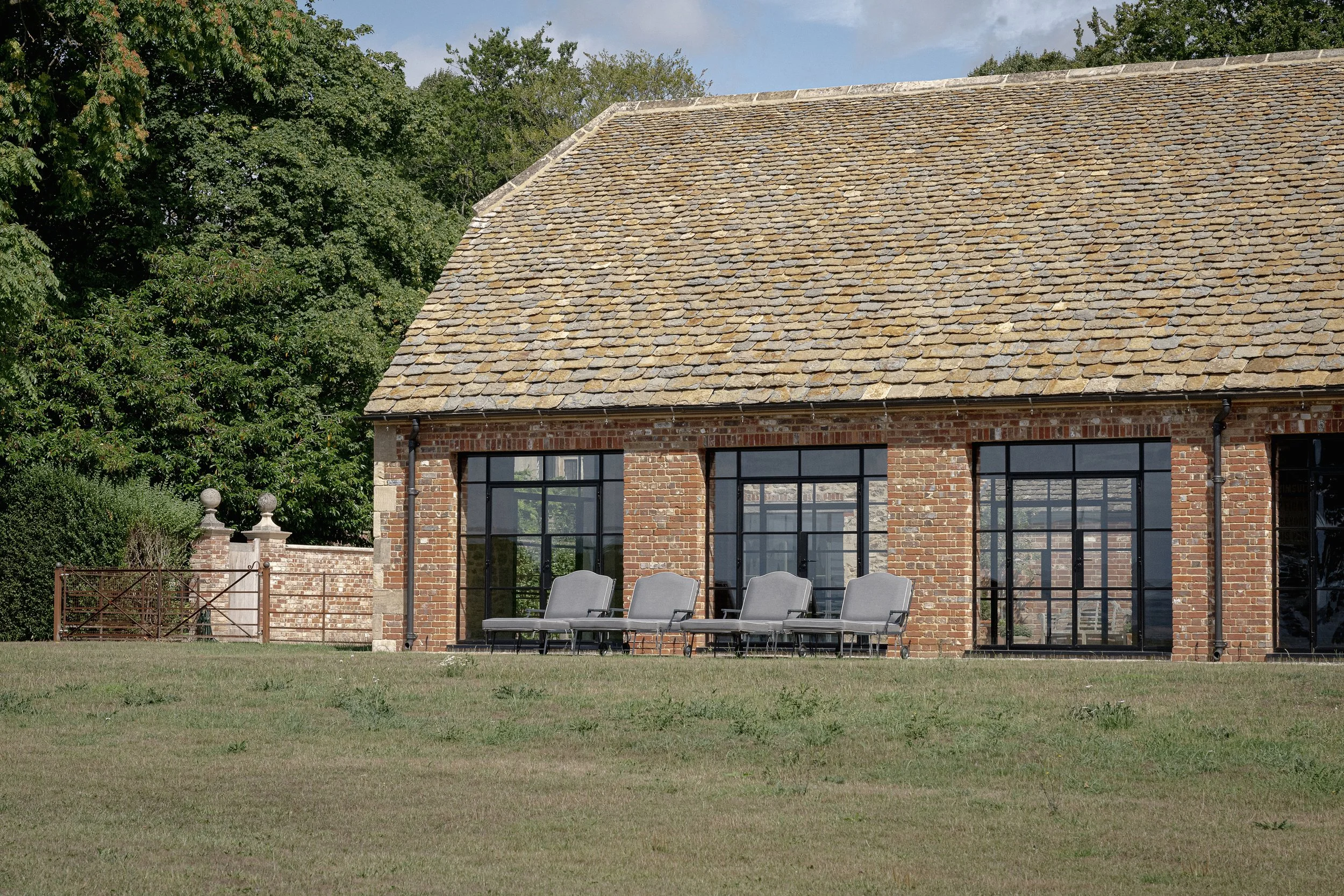 A brick building with large black-framed windows and a sloped, tiled roof, situated on a grassy lawn with trees in the background.