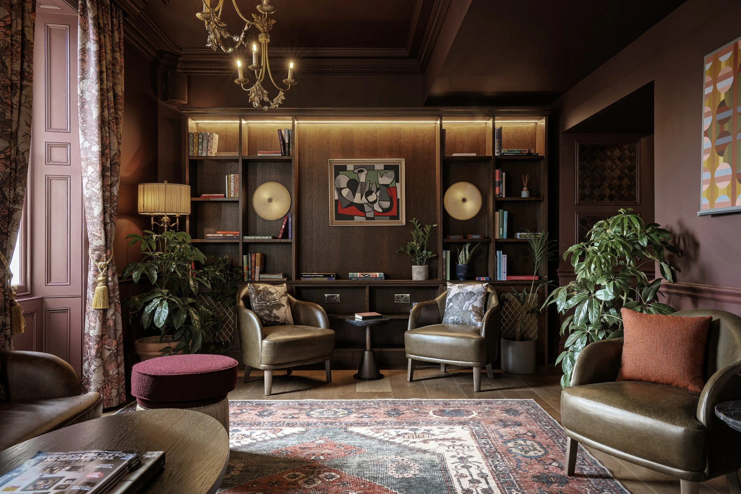 A cozy living room with dark walls, wooden shelves with books, a framed abstract artwork, and plants. It has leather chairs, a patterned rug, and warm lighting from a chandelier and lamps.