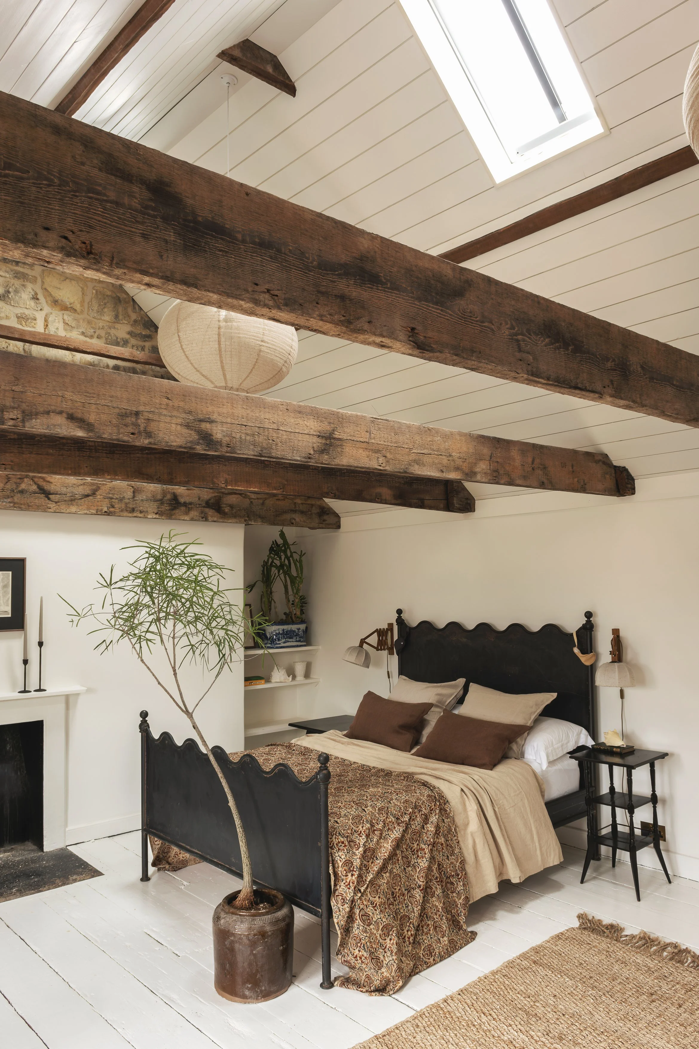 A bedroom with a black bed frame, beige bedding, and brown pillows, a white fireplace, green plants, a small black side table, and a large potted plant. Exposed wooden ceiling beams and a skylight ceiling are visible.