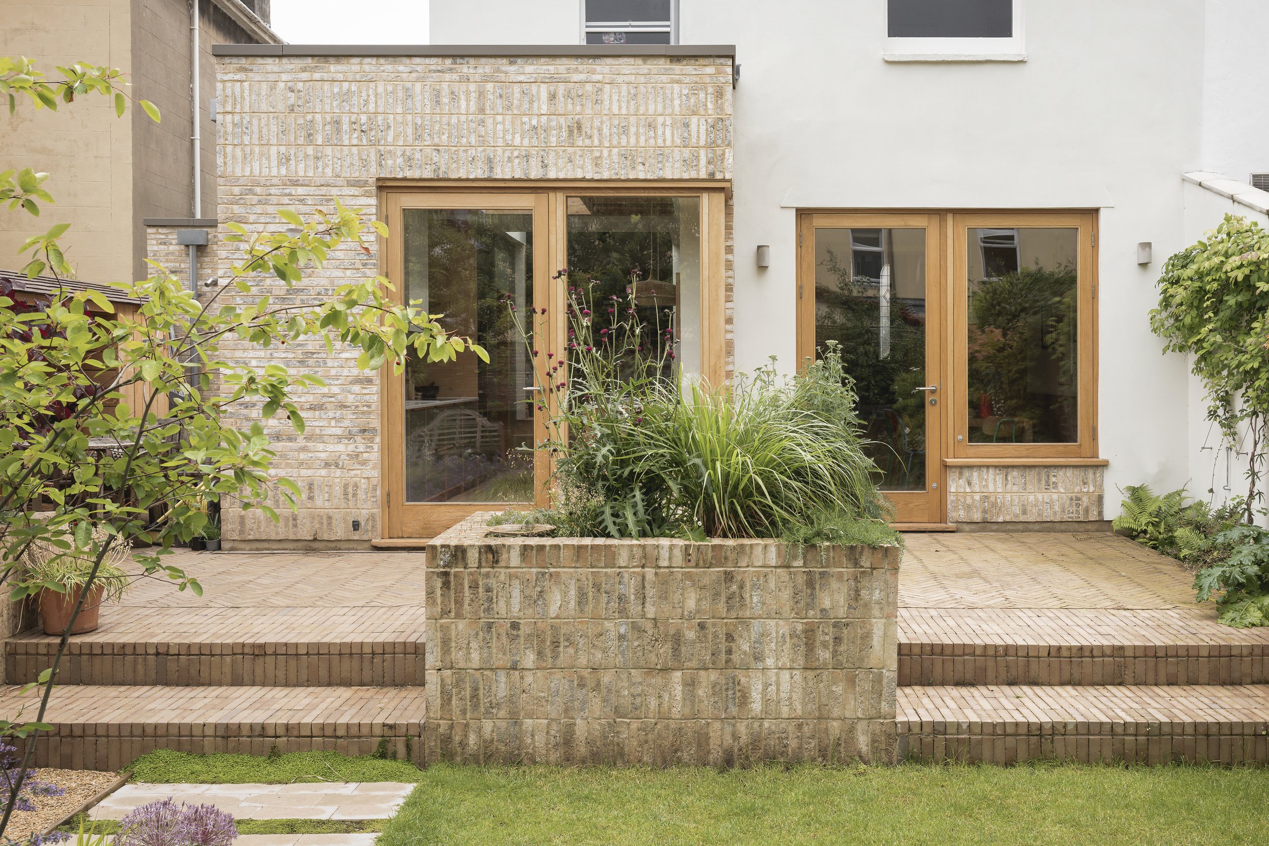 Modern house backyard with brick steps, a raised brick garden bed with green plants, and two large glass doors with wooden frames leading inside. There are trees and shrubs around, and a well-maintained lawn in the foreground.