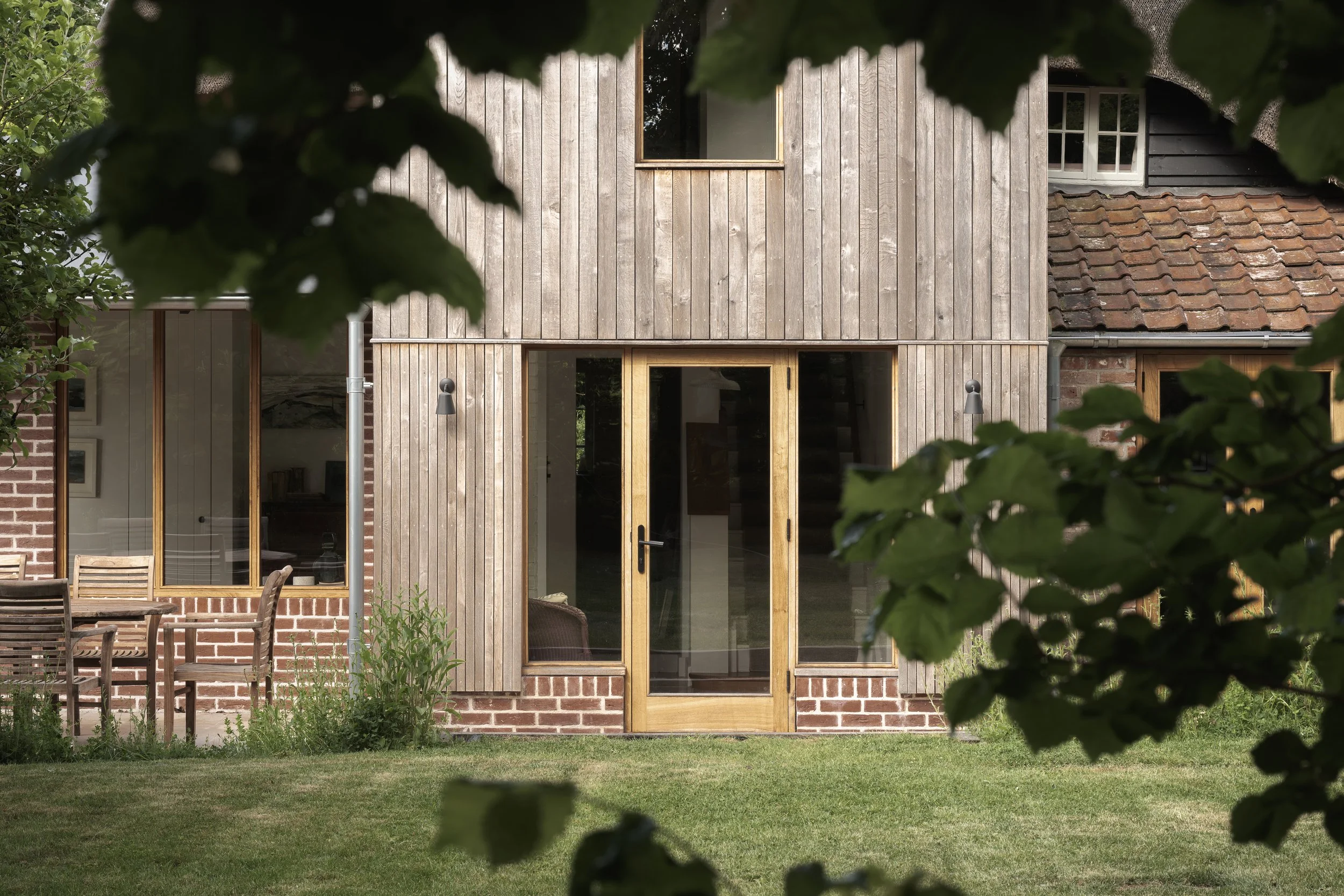 View of a modern house with wooden and brick exterior, featuring large glass doors and windows, surrounded by green grass and plants, with tree leaves in the foreground.