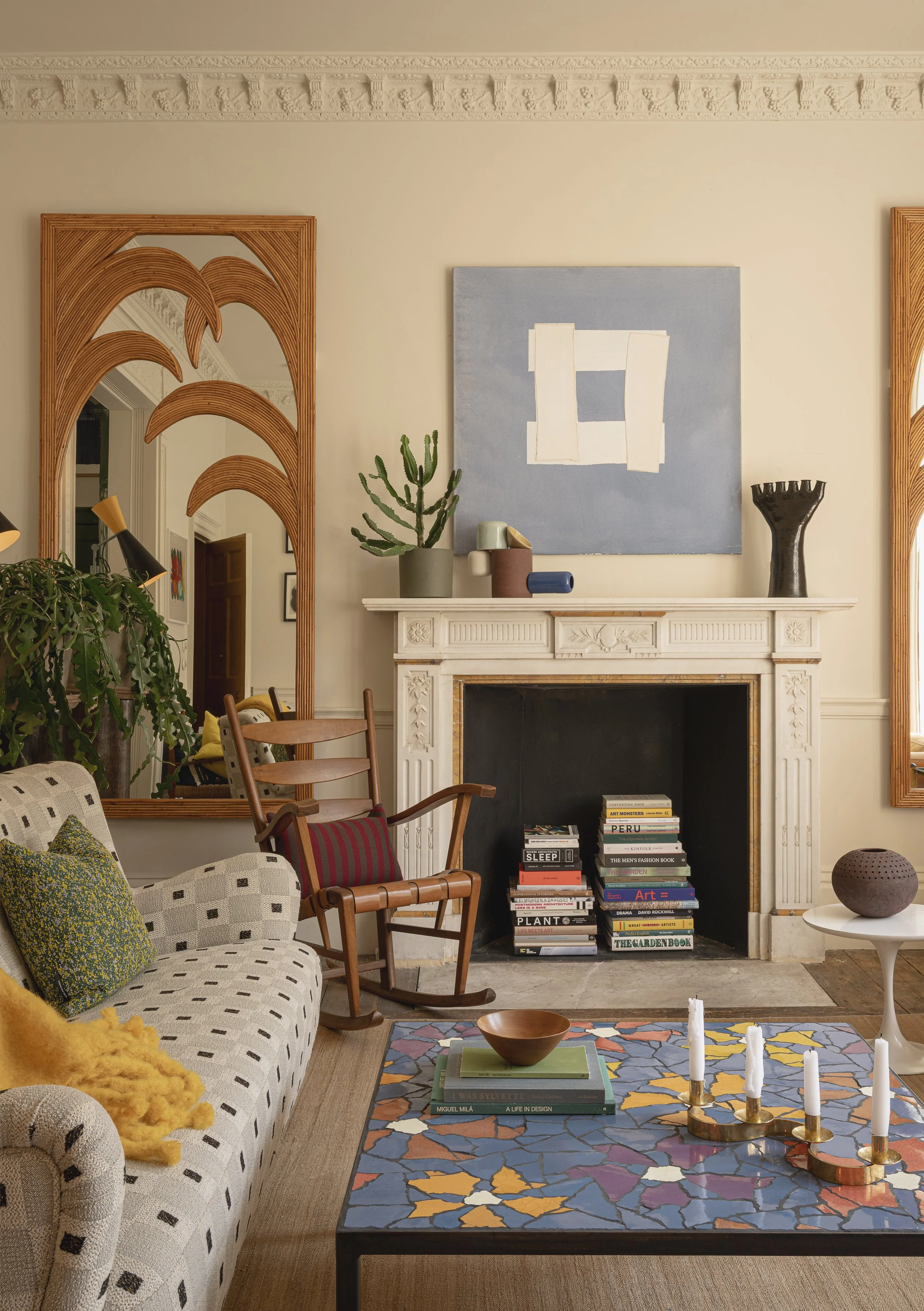 Decorative living room with a patterned sofa, wooden armchair, colorful rug, modern coffee table with books and a bowl, stacked books inside a fireplace, artwork above the fireplace, green cactus plant on mantel, large mirror on wall, and candles on 