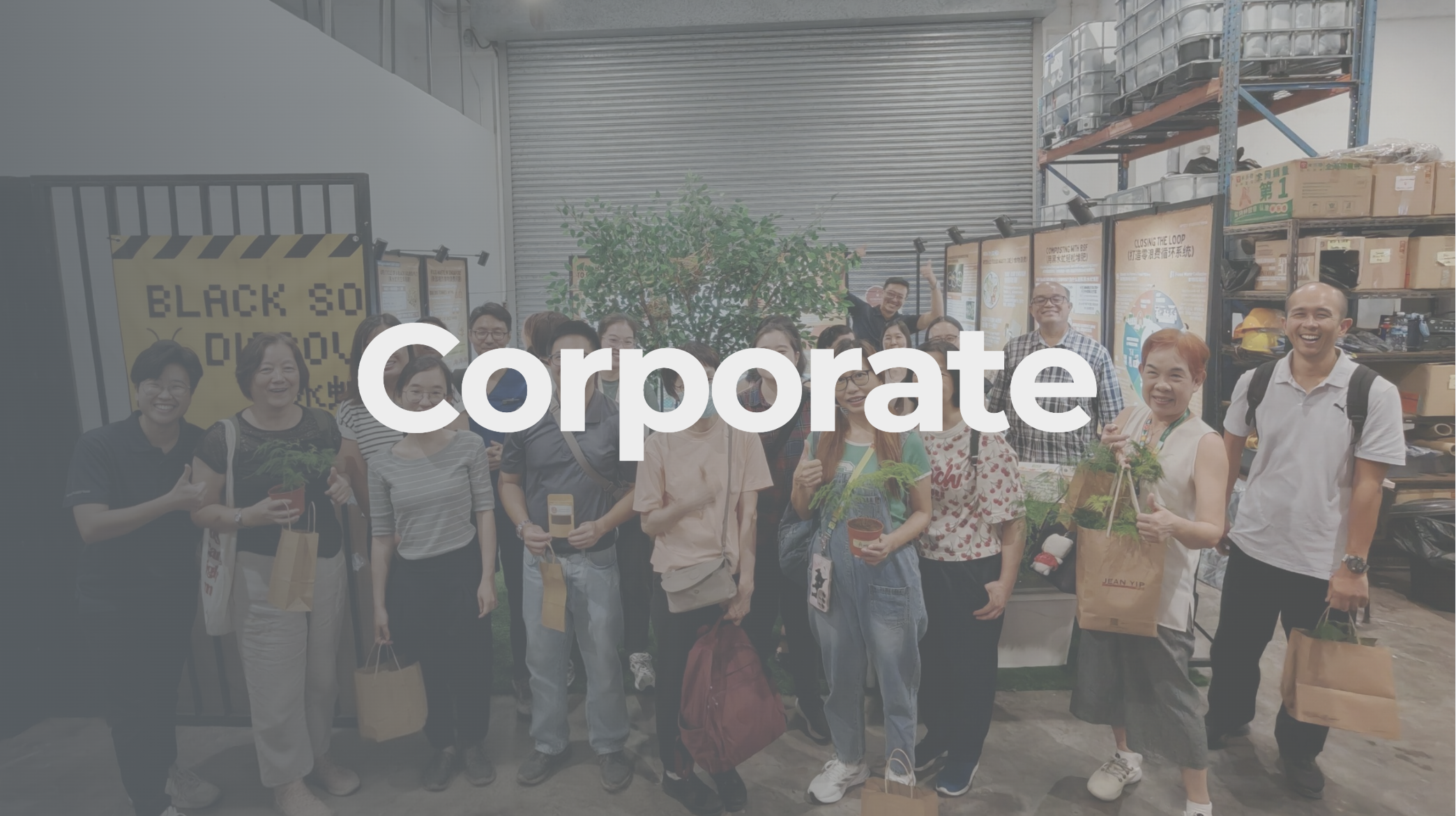 Group of diverse people standing in warehouse or workshop, smiling and holding potted plants and gift bags, with shelving and boxes in background.