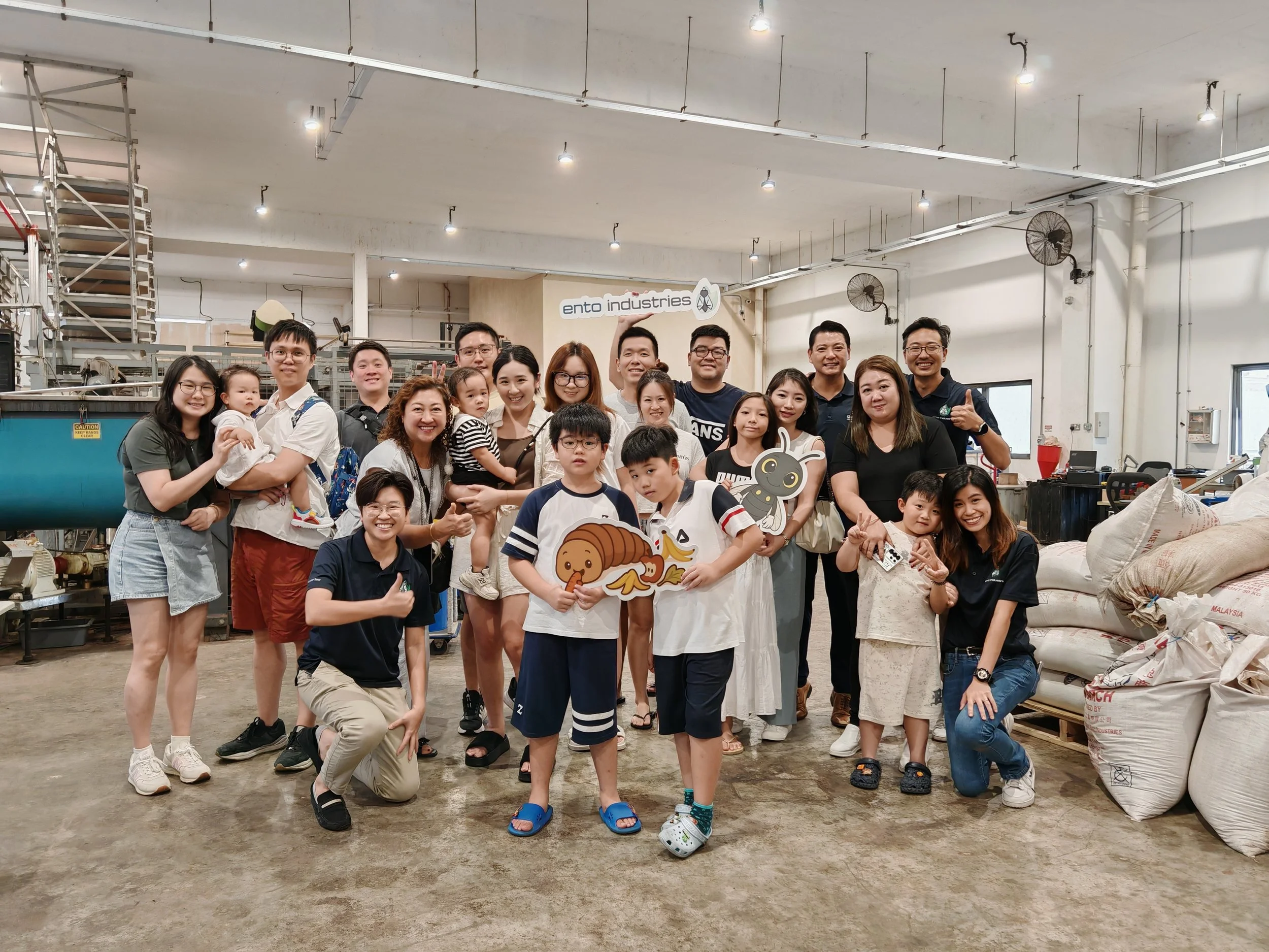 Group of adults and children smiling and posing for a photo inside  Ento Industries' factory with machinery and large sacks of materials.