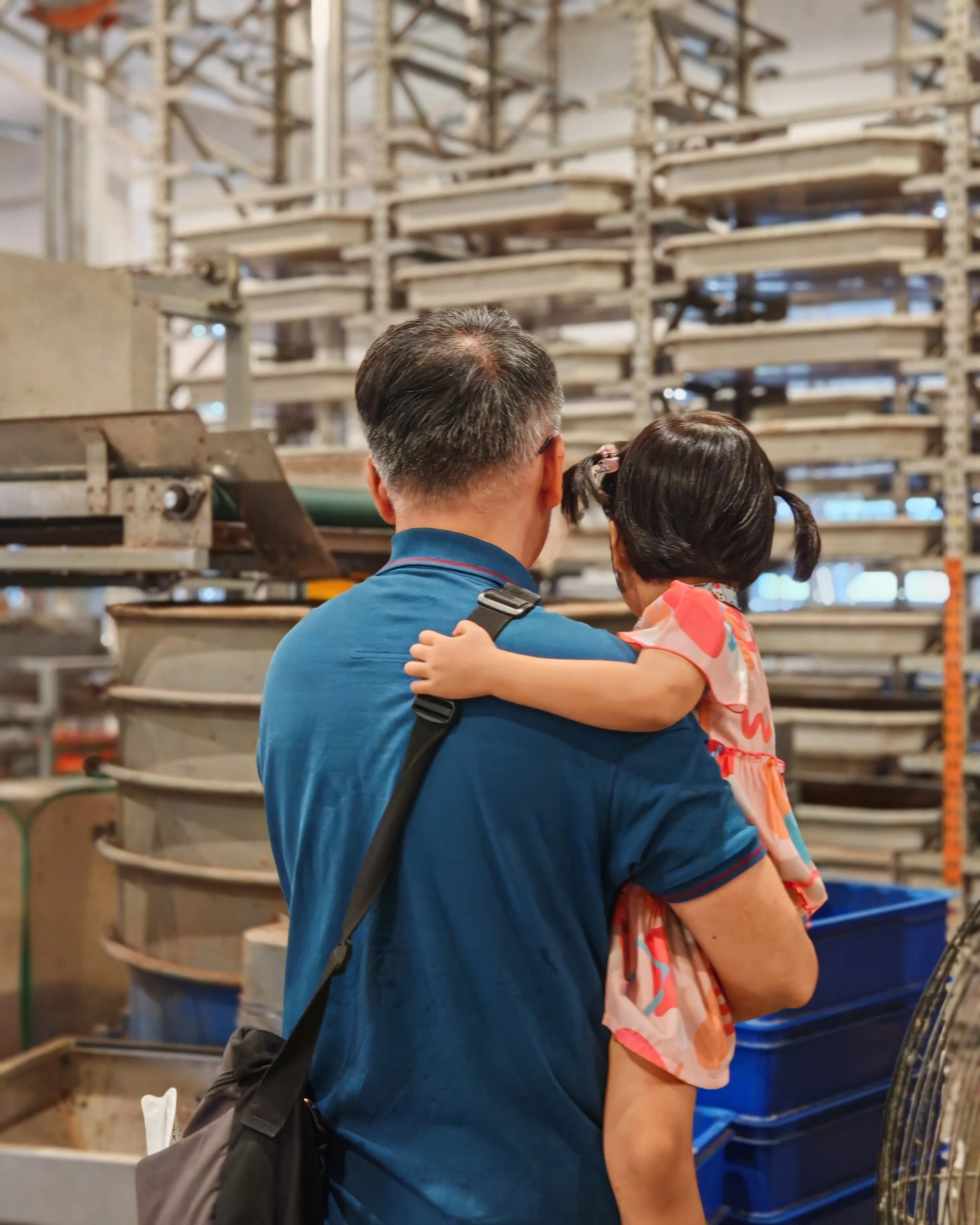 A man and a young girl are shopping in a store, standing close together with the man holding the girl in his arms. The background shows shelves with empty trays and metal carts.