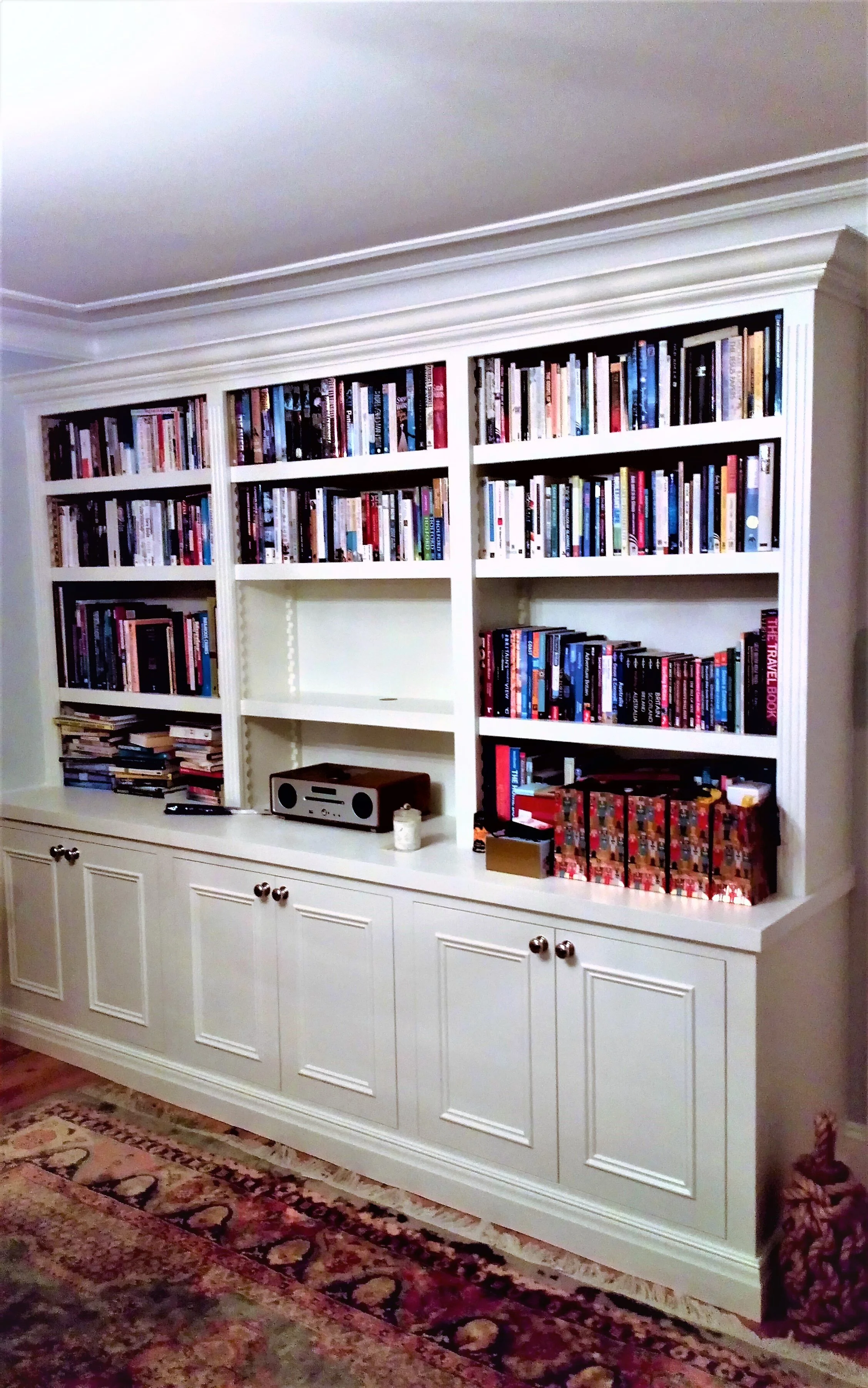 White built-in bookshelf filled with books, with electronic device, candle, and decorative boxes on the bottom shelf. A patterned rug is partially visible on the floor.