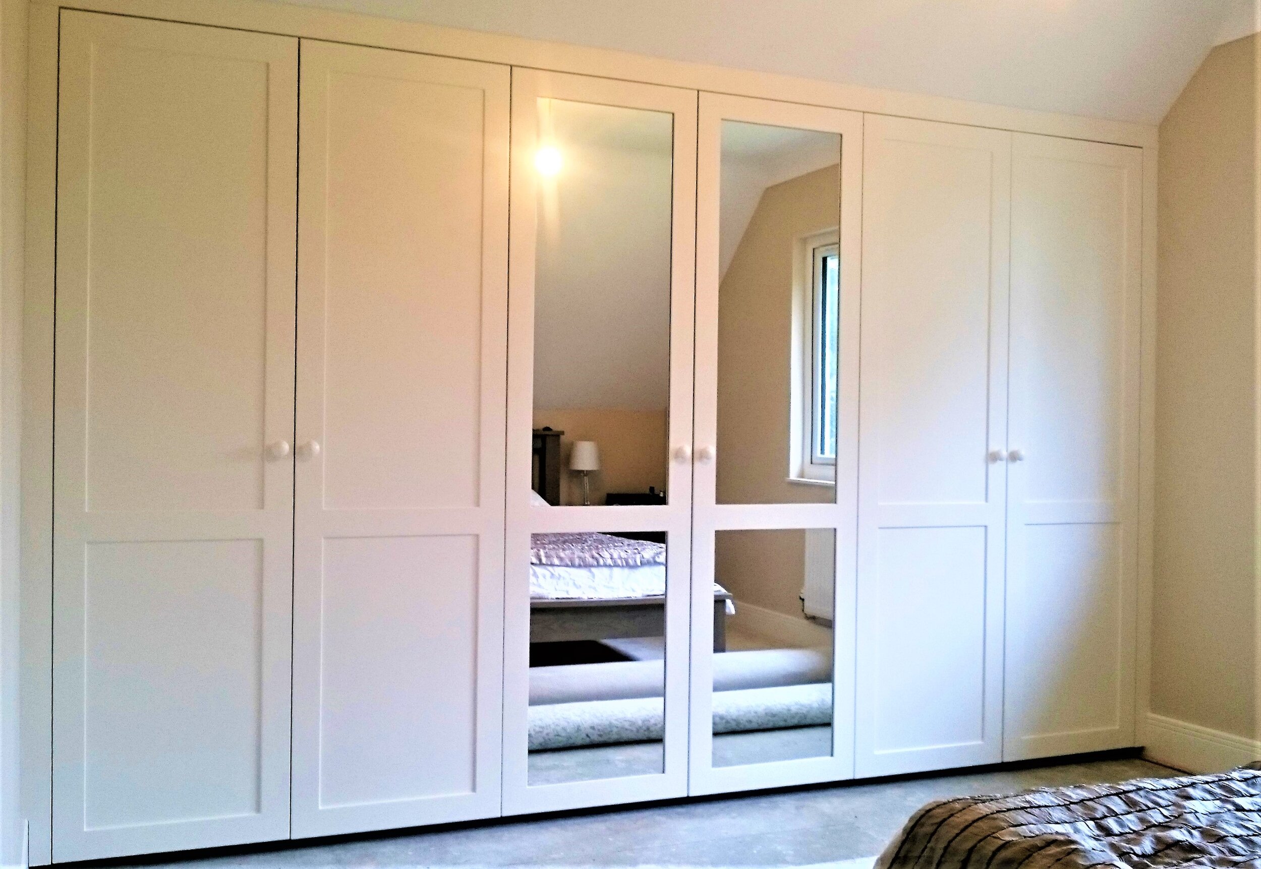 White wardrobe with four doors, three with panels and one with mirrors, located in a bedroom with beige walls and a window. The reflection shows a bed with a blanket and a lamp on a nightstand.