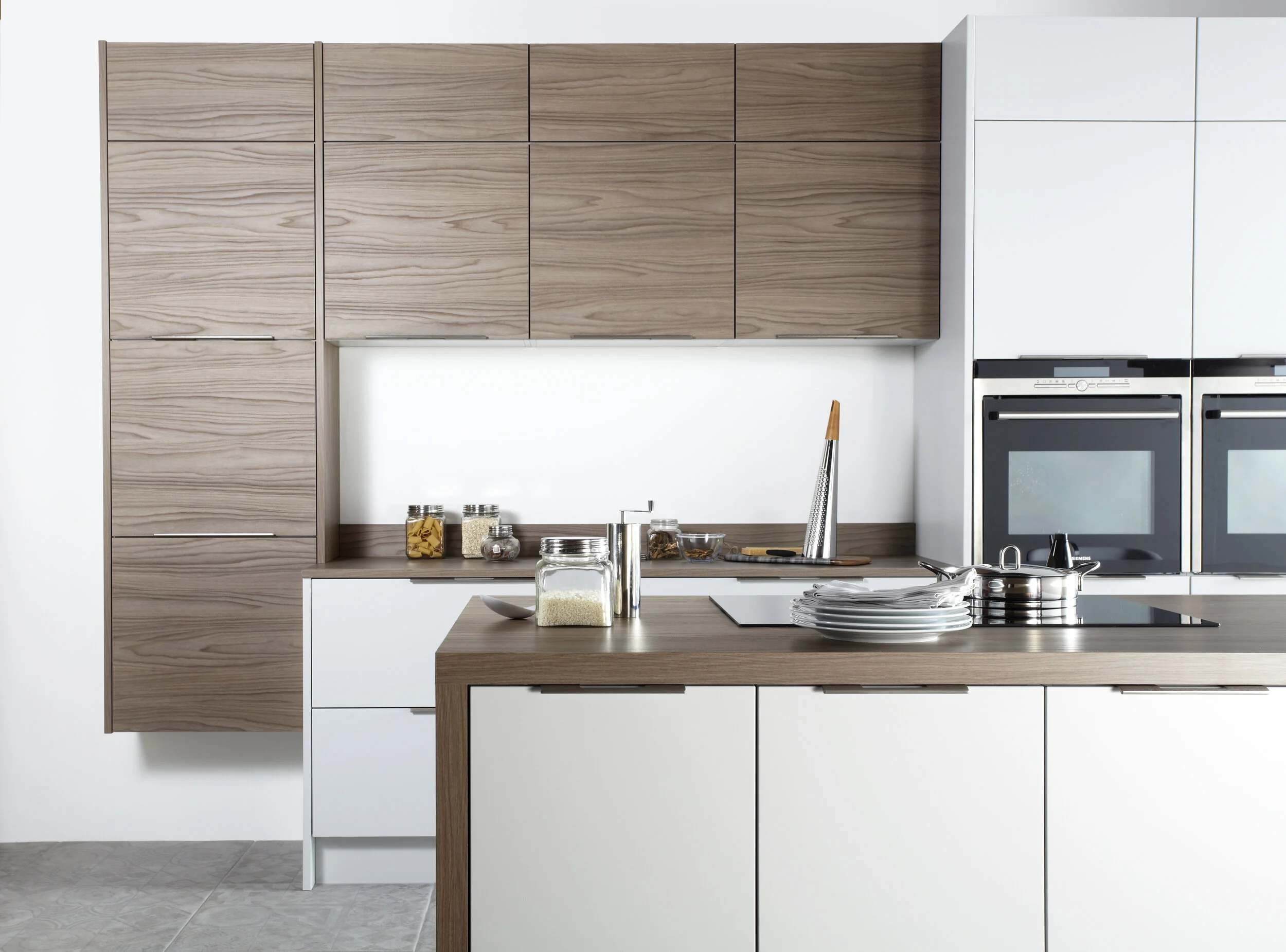 Modern kitchen with wooden and white cabinets, countertop with jars of pasta, rice, and spices, a sink, cutting board with a grater, and stacked plates.