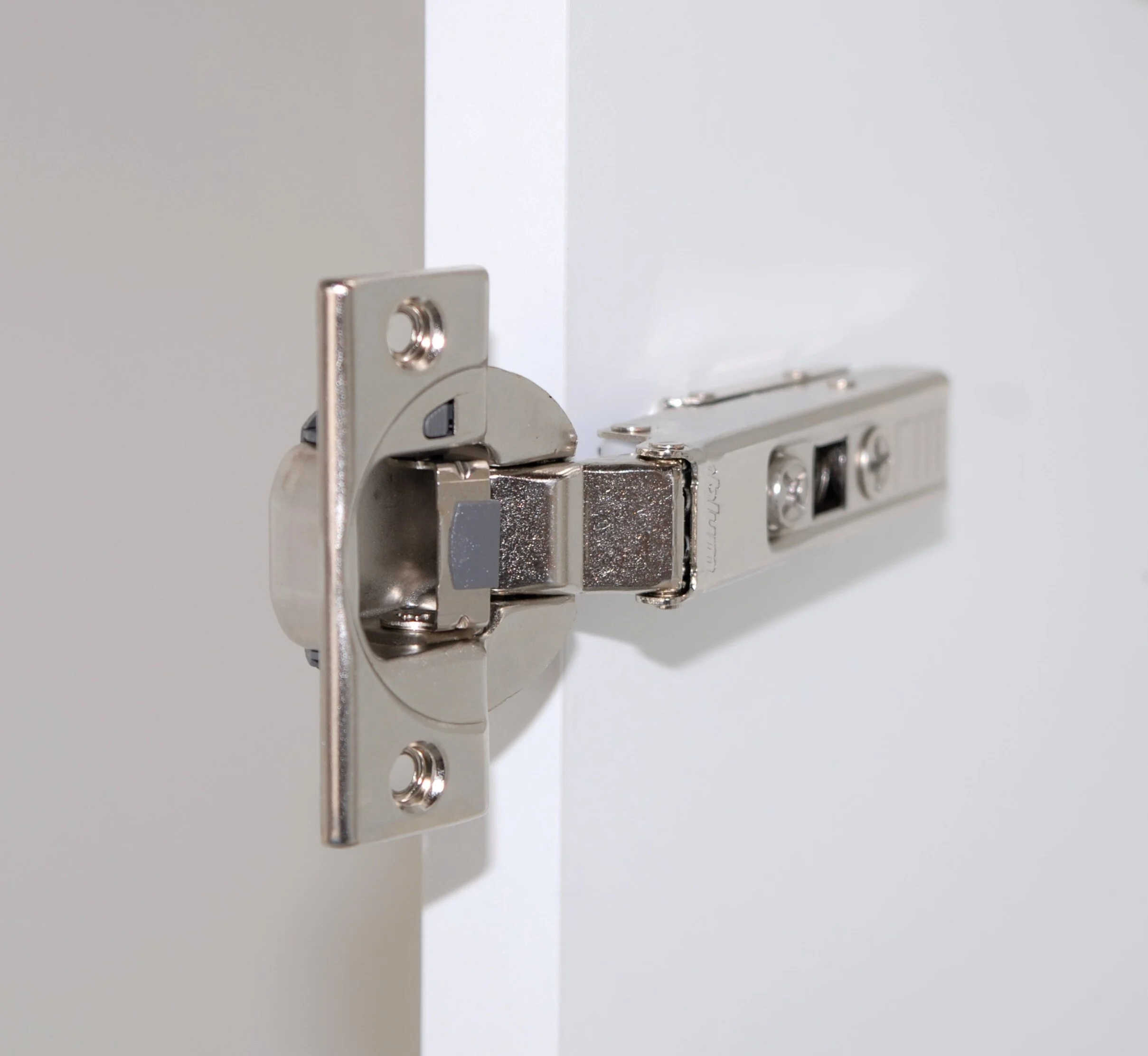 Close-up of a metal cabinet door hinge installed on a white cabinet.