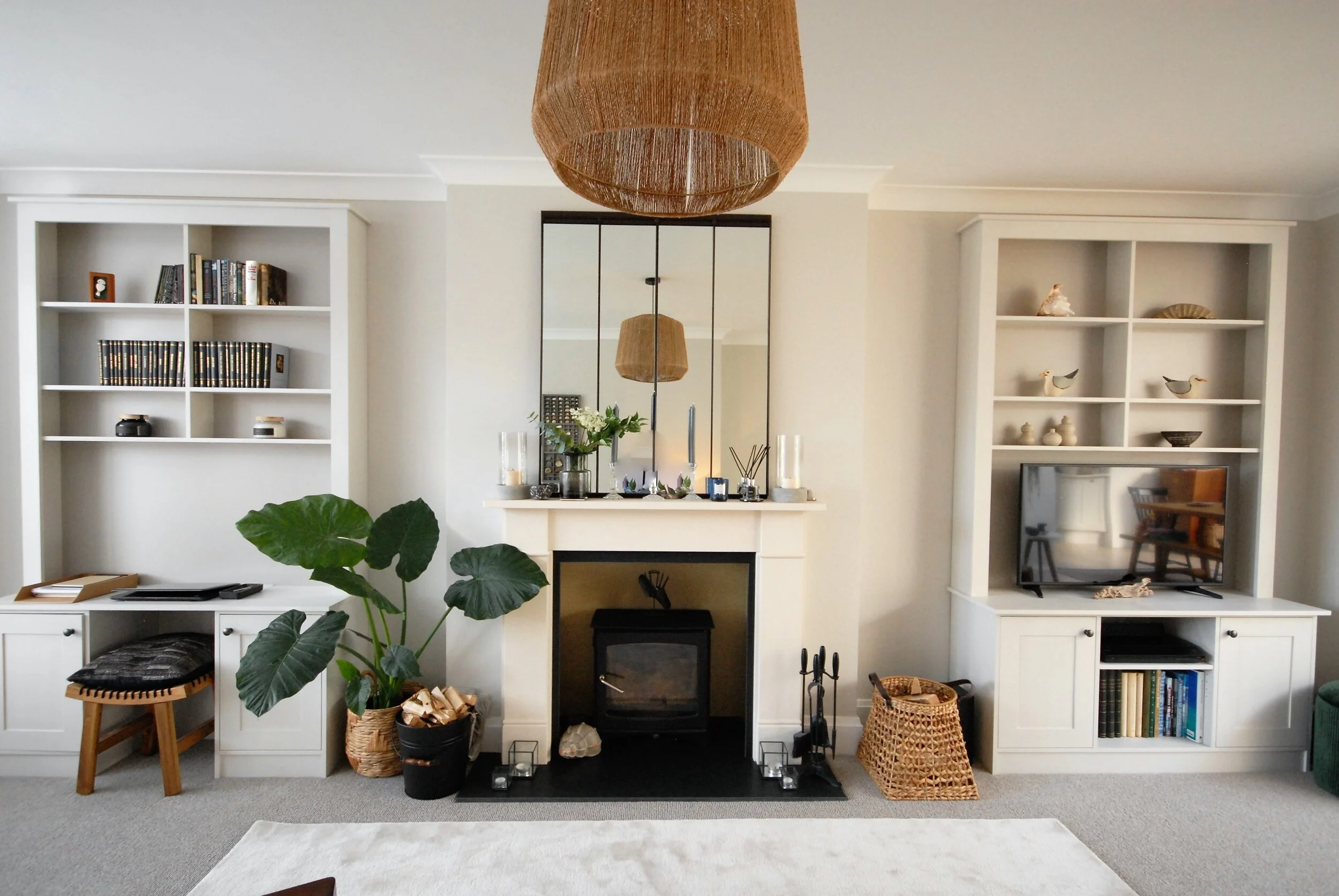 Living room with white built-in shelves on each side of a fireplace, a large mirror above the fireplace, a large potted plant on the left, a wicker basket and fireplace tools on the right, and a flat-screen TV on the right shelf. Decor includes books