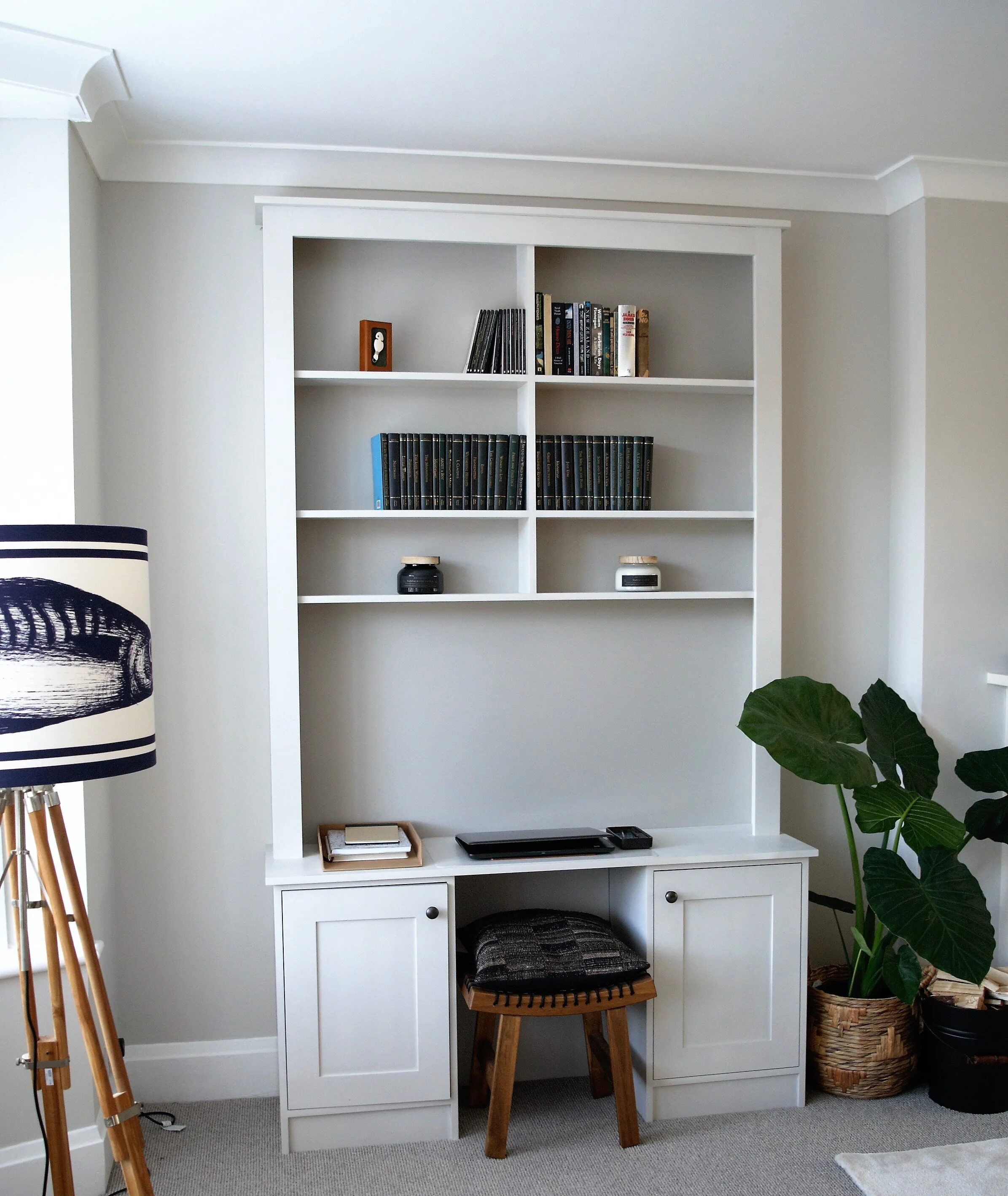 White built-in bookshelf with books and decorative items, a small wooden stool with a seat cushion, a potted plant, and a floor lamp with a patterned shade in a room.
