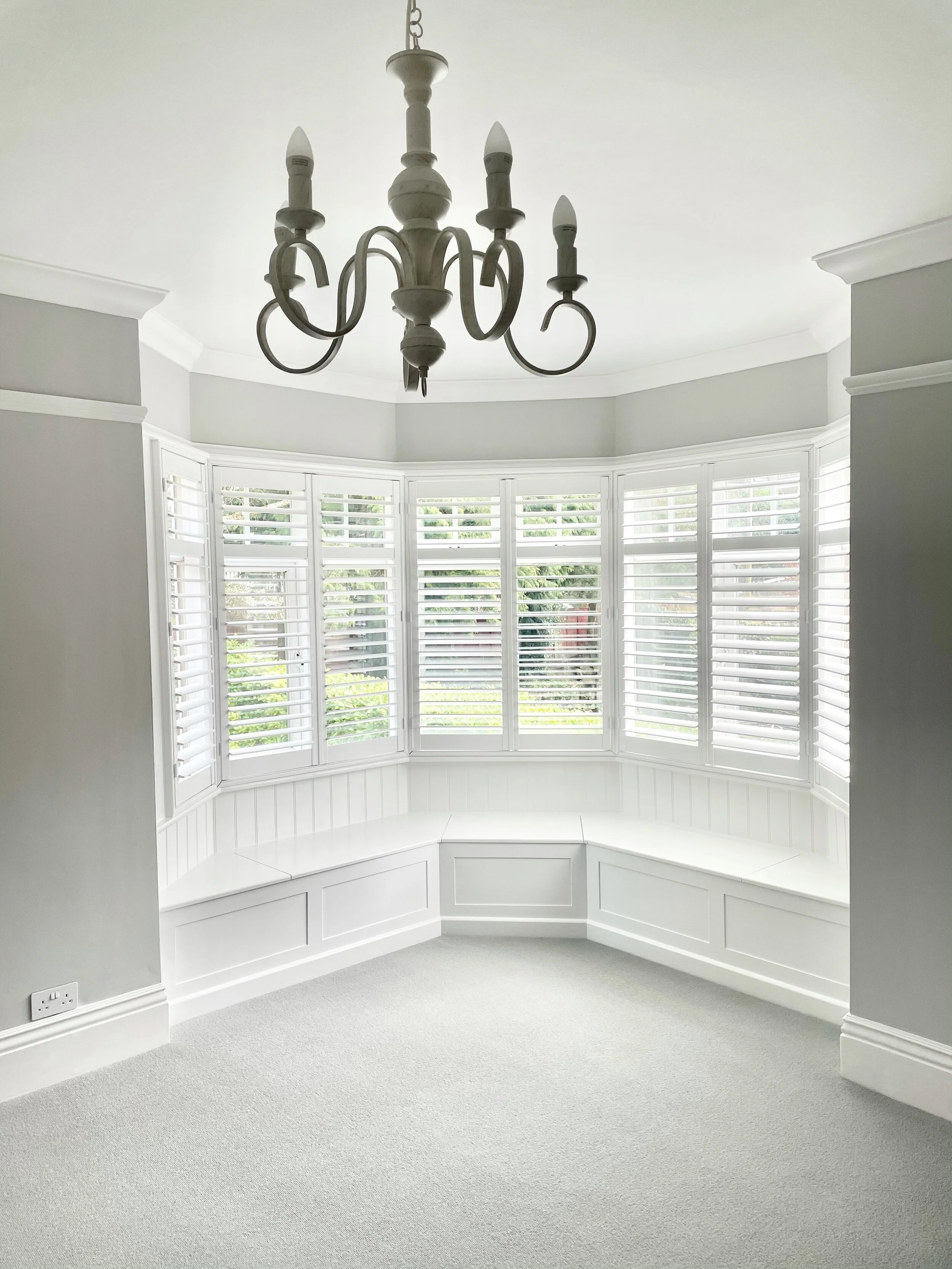 Bright room with a large bay window with white shutters, built-in window seat, and a white chandelier hanging from the ceiling.