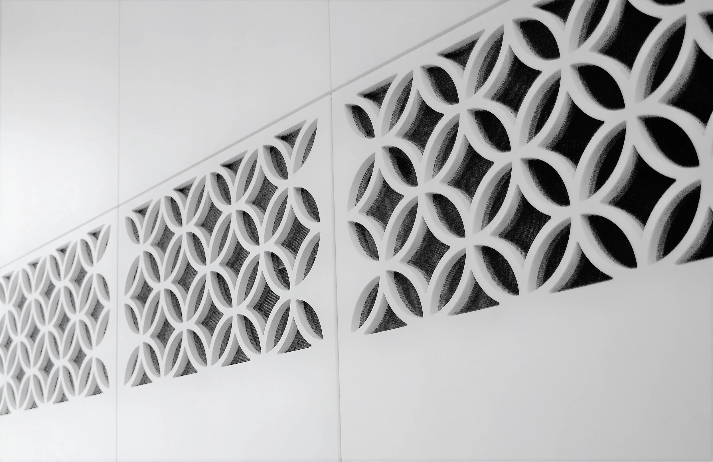 Close-up of a white decorative wall panel with a repeating geometric pattern of circular and petal-shaped cutouts, set against a plain white wall.