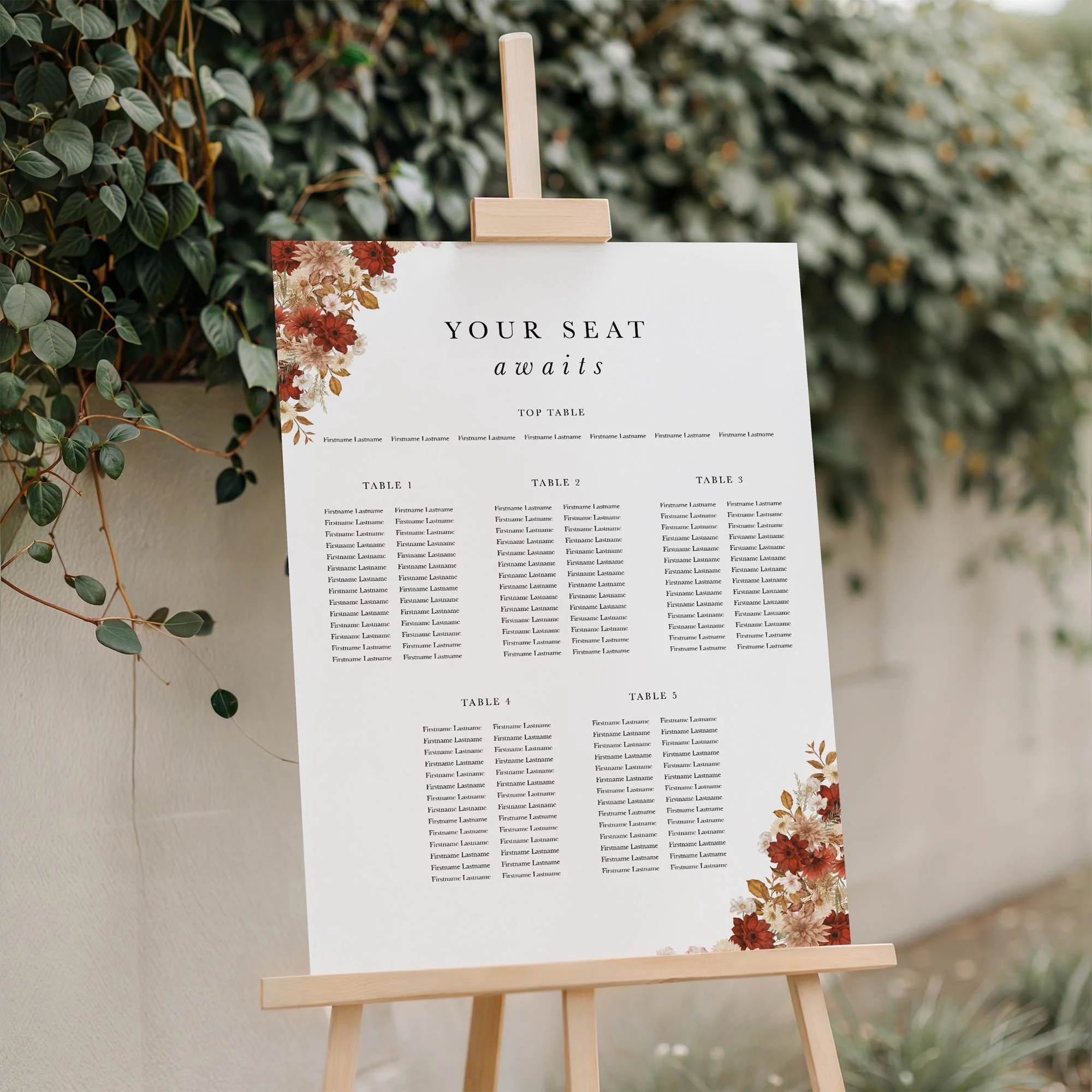 Rustic wedding seating plan displayed on a wooden easel, featuring a bold crimson and cream autumn floral design with multiple guest tables