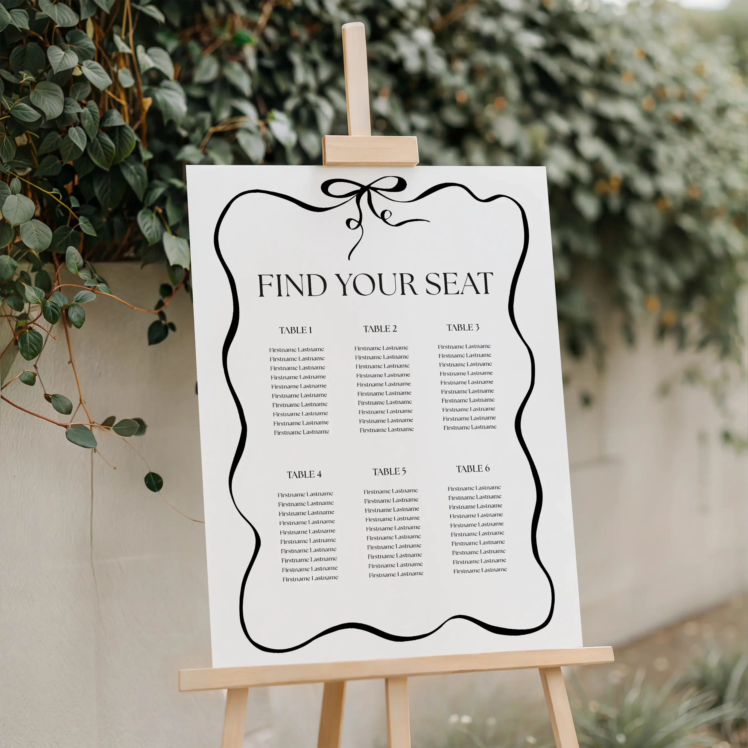 Black and white wedding seating plan with bow motif and wavy border displayed on a wooden easel with greenery background — Black Tie collection by Wonder Wedding Stationery