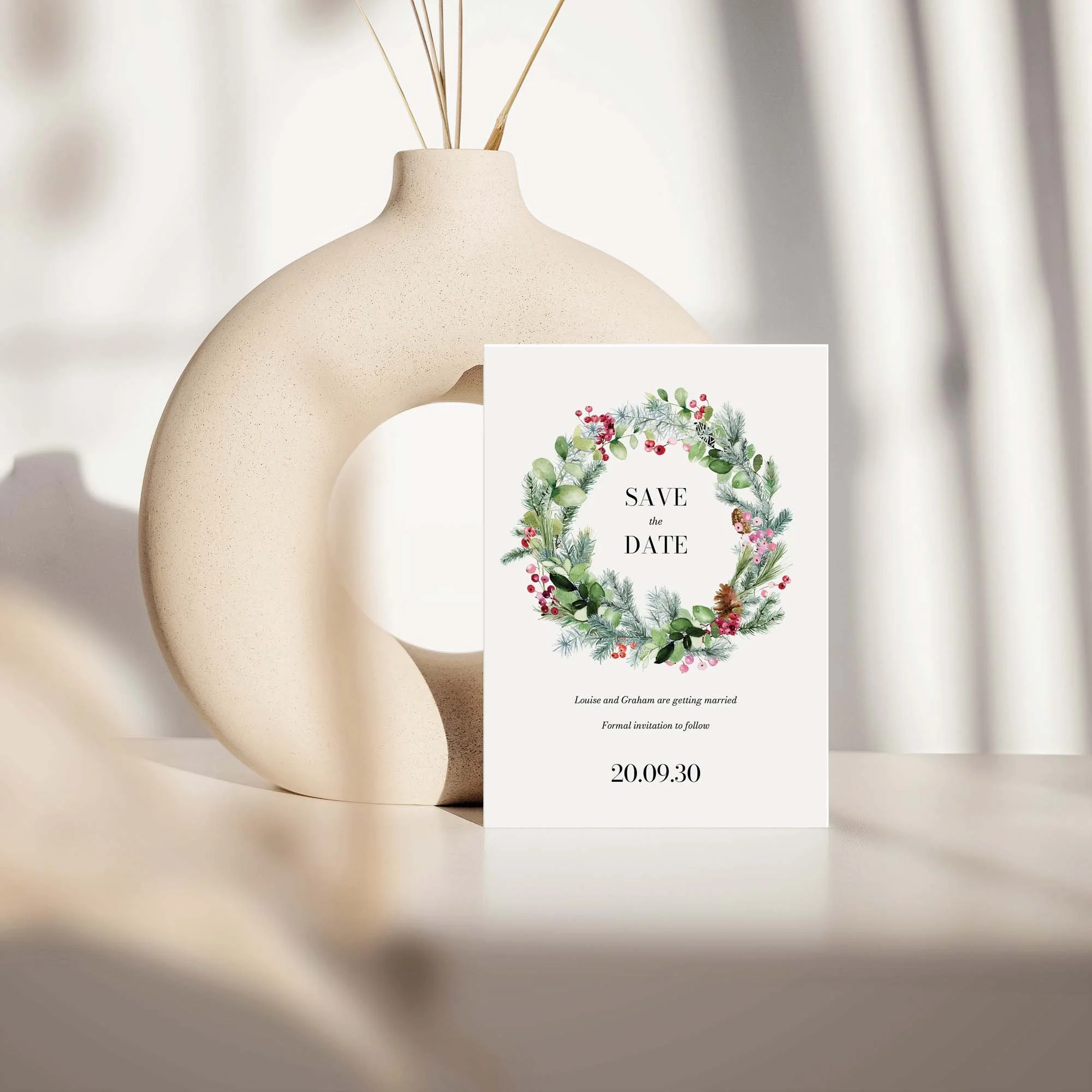 Festive save the date card with holly and red berry Christmas design displayed against a cream ceramic vase on a neutral background