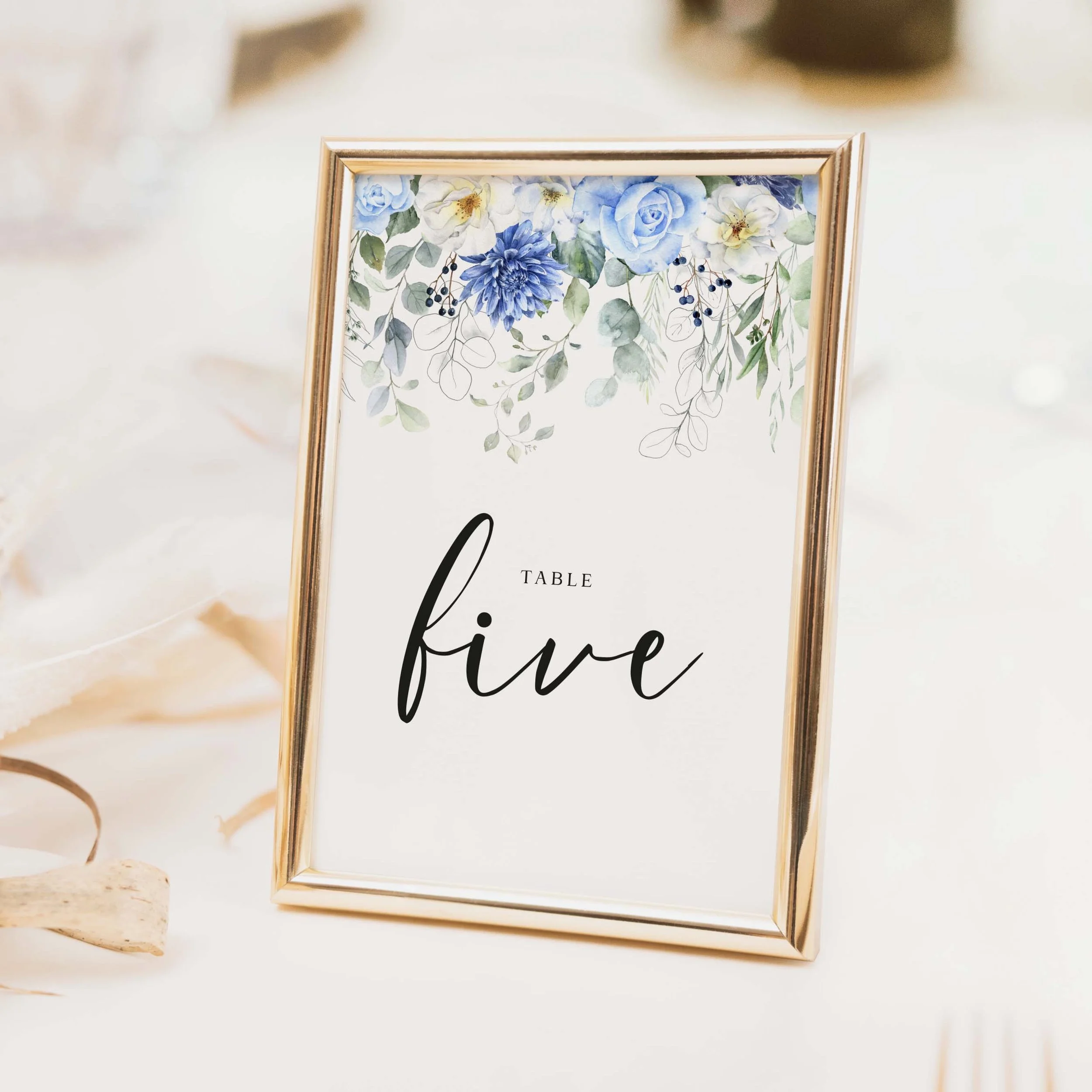 Blue Floral wedding table number with abundant blue and white floral arrangement, soft green foliage and flowing script typography printed on luxury off-white textured card