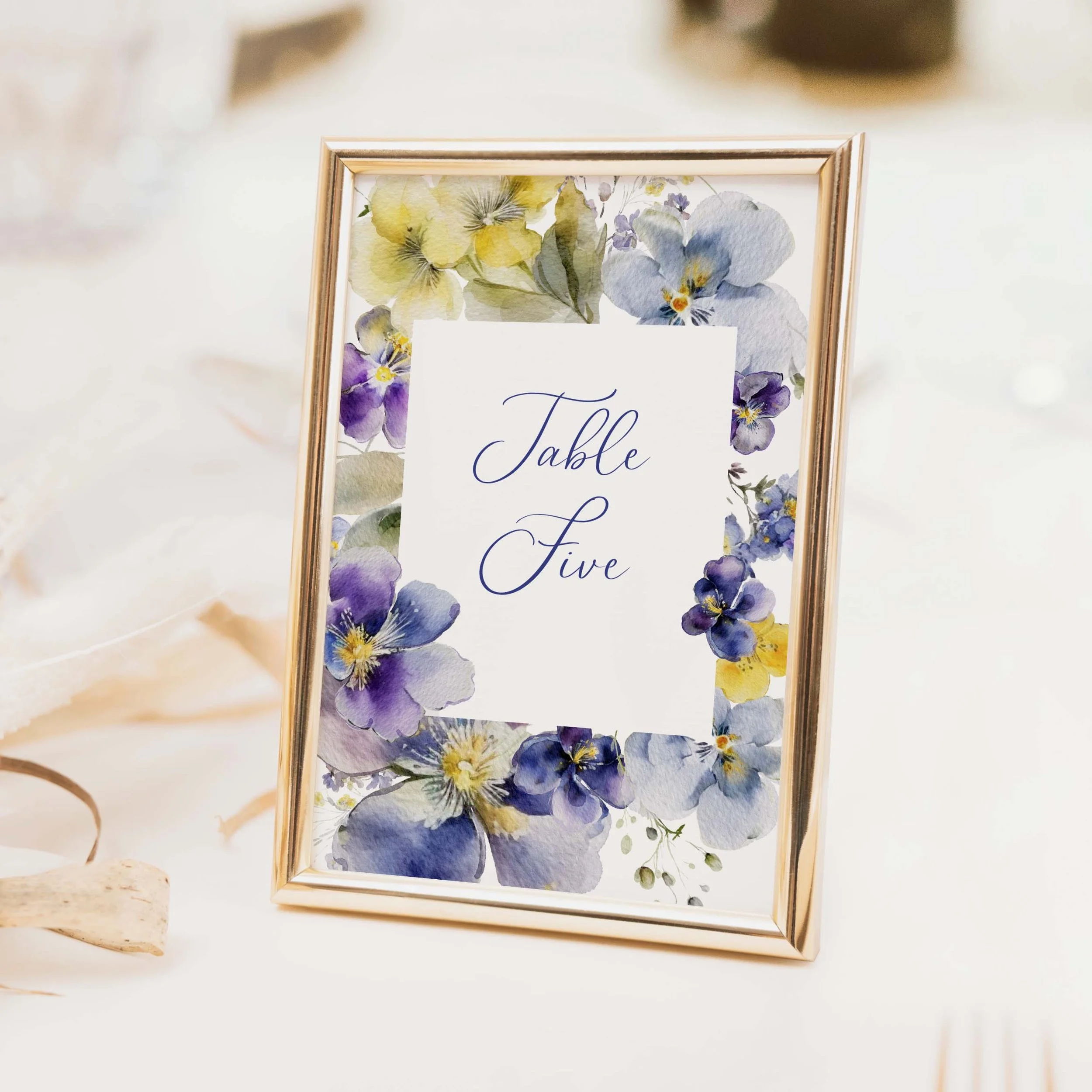 Purple and yellow botanical floral wedding table number with watercolour violet arrangement and flowing script typography on luxury off-white card