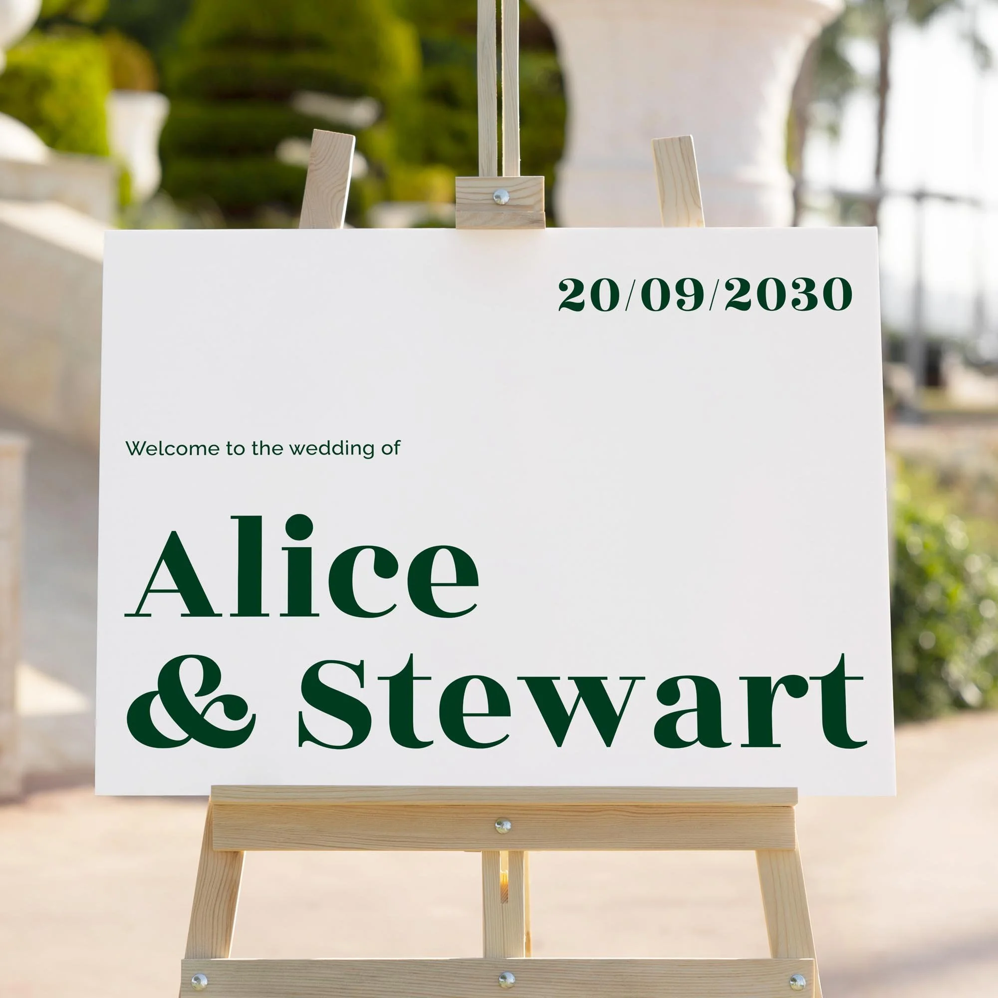 Elegant modern wedding signage with serif font in deep forest green, displayed on a wooden easel with greenery and stone columns in the background.