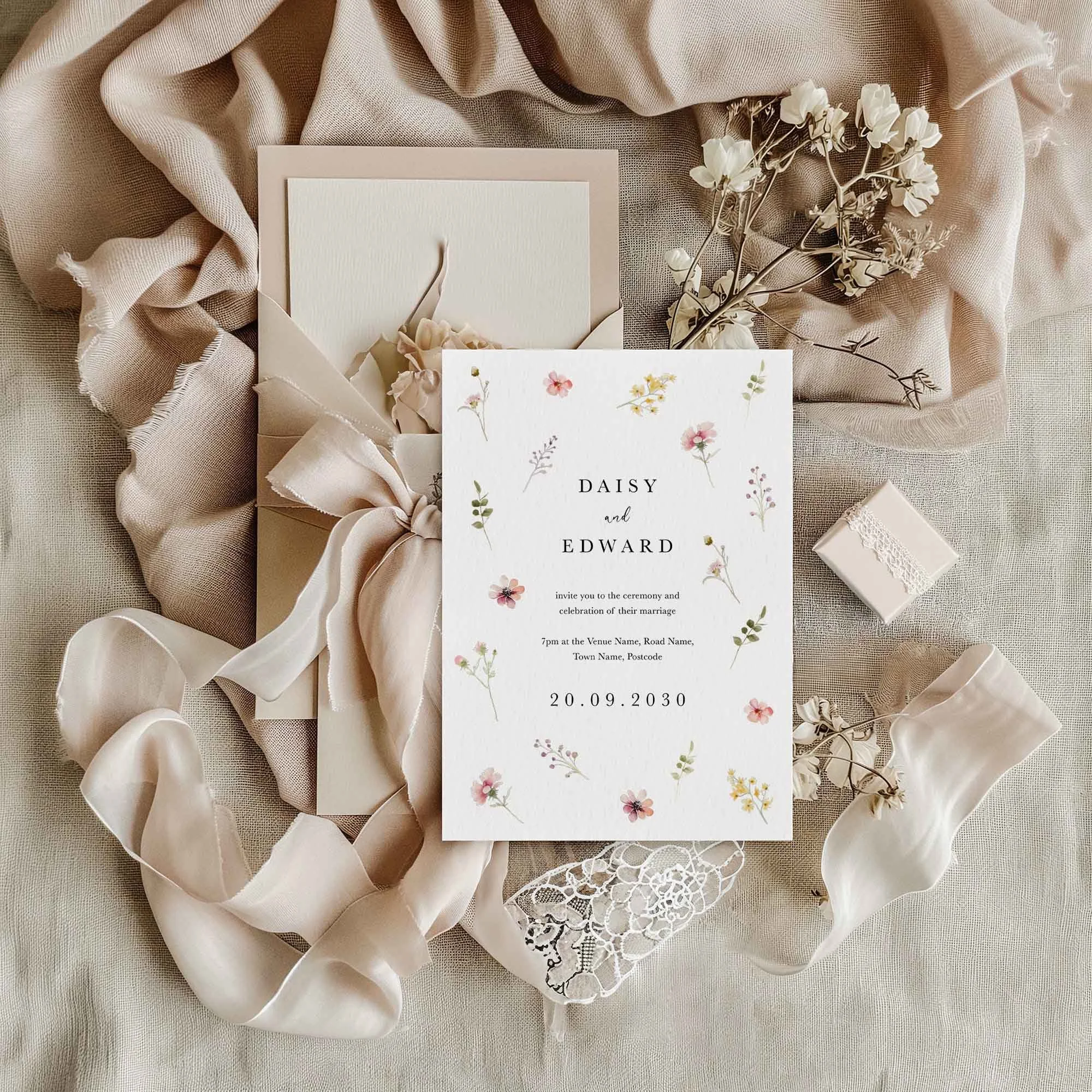 Flora wedding invitation flat lay featuring scattered pastel wildflowers, displayed on sage linen fabric with dried flowers and a small gift box